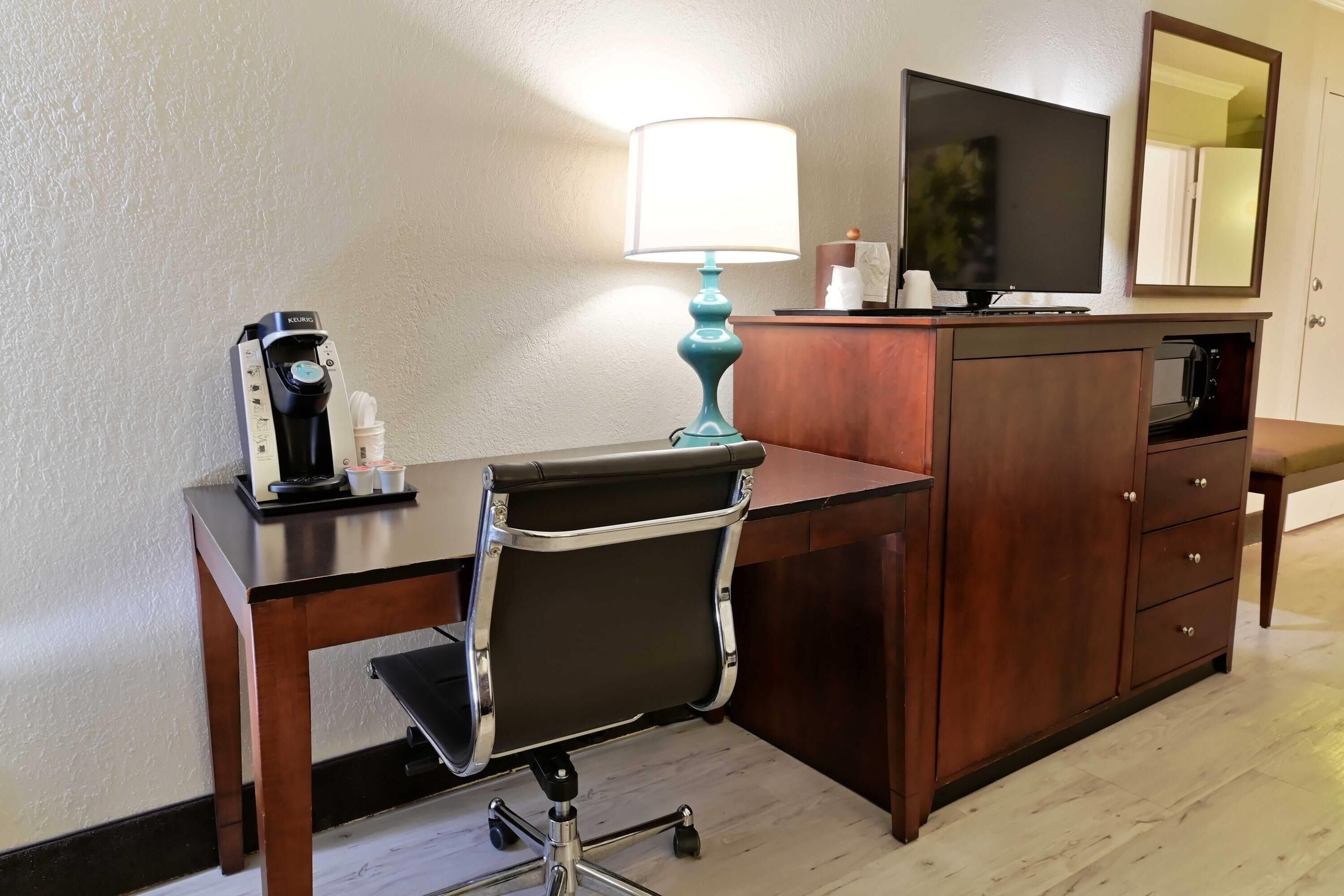 A hotel room with a wooden desk, a black office chair, a teal desk lamp, a coffee maker, a television, a mirror, and a wooden cabinet.