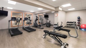 Ein Fitnessstudio mit Laufbändern, Crosstrainern, einer Langhantelbank und einem Fernseher an der Wand.