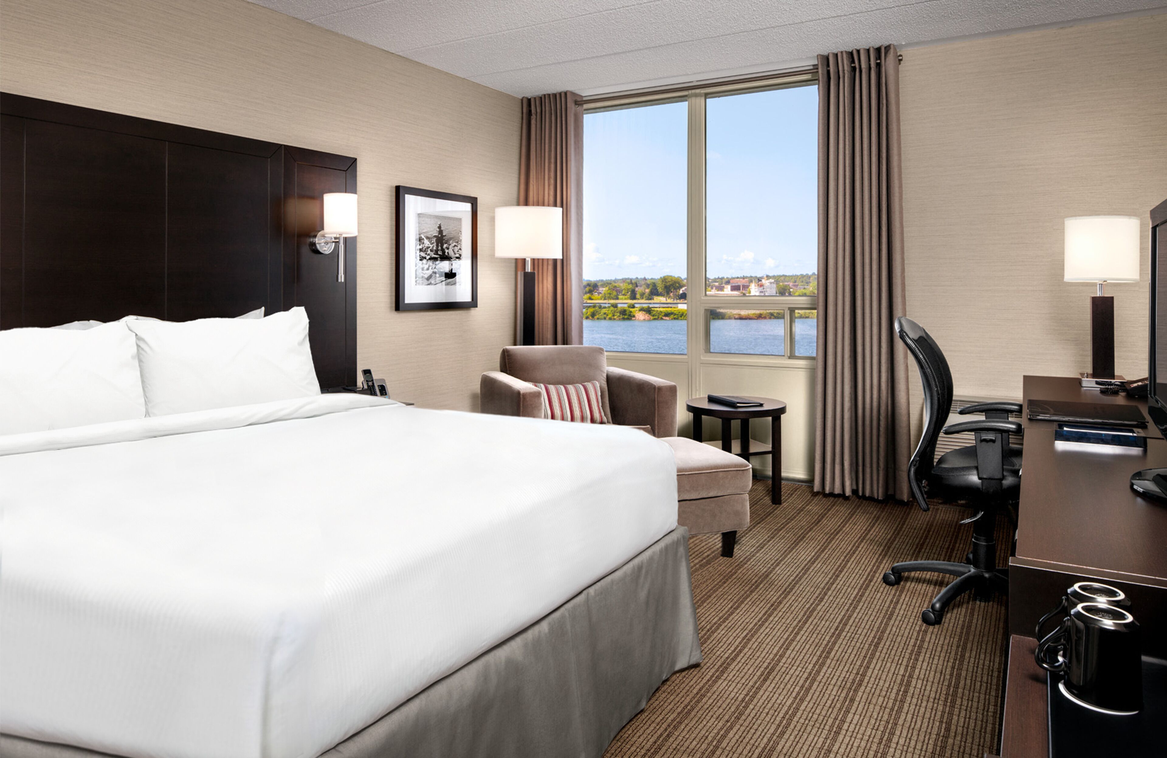 A hotel room with a large bed, a desk, a chair, and a view of a body of water through the window.