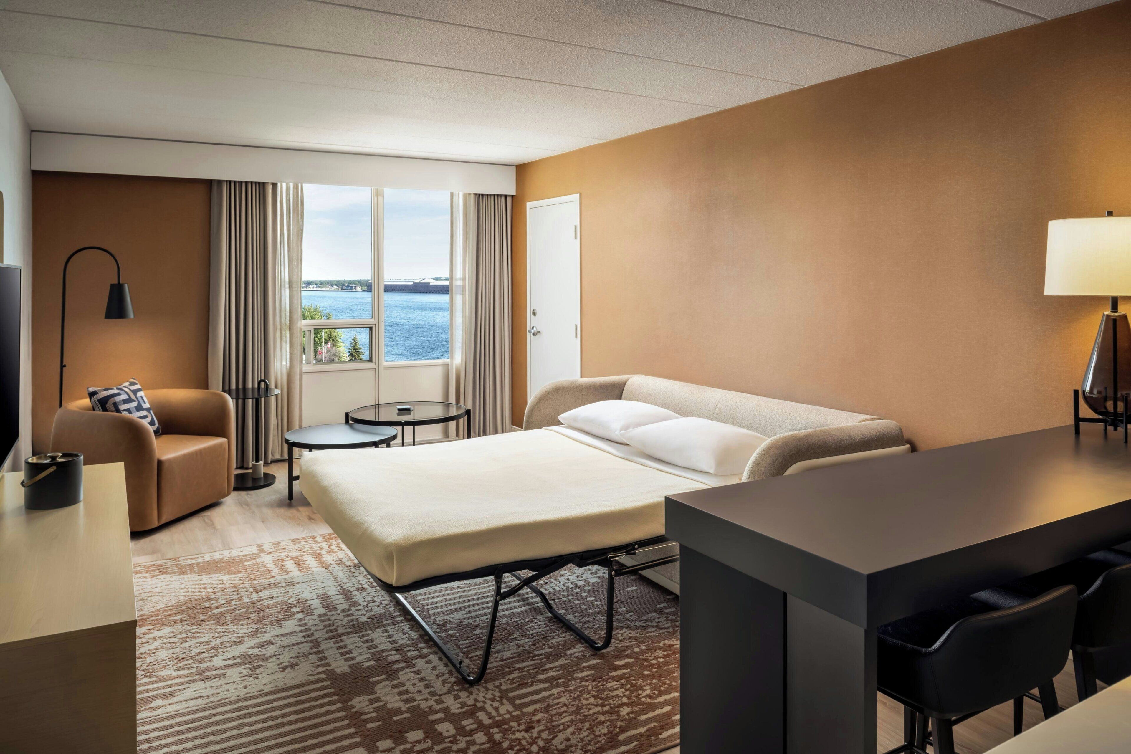 A hotel room with a bed, a desk, a chair, a small table, a lamp, and a view of the water.