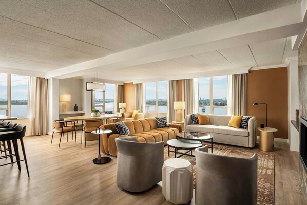 A spacious living room with a large window, a sofa set, a dining area, and a view of the water.