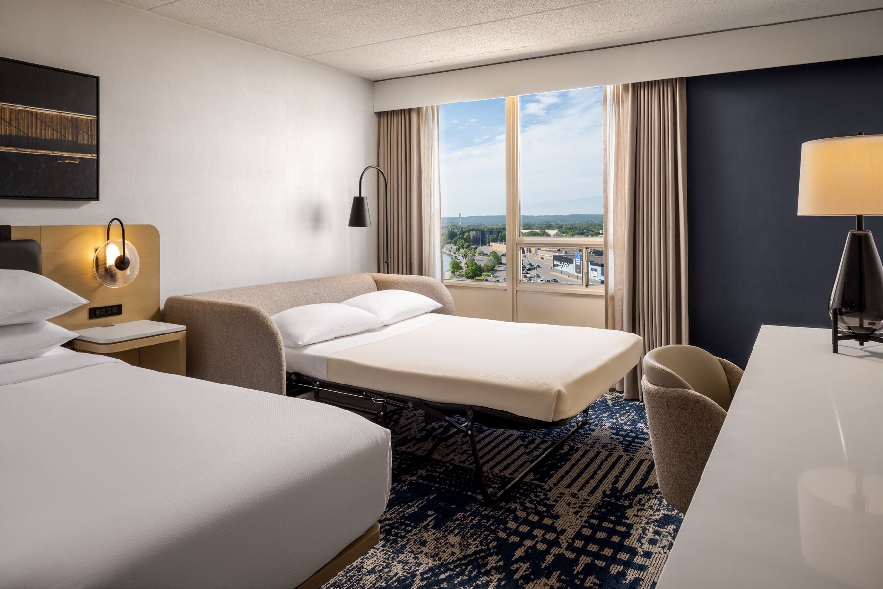 Habitación de hotel con dos camas, un escritorio, una silla, una lámpara y vistas a un paisaje urbano.
