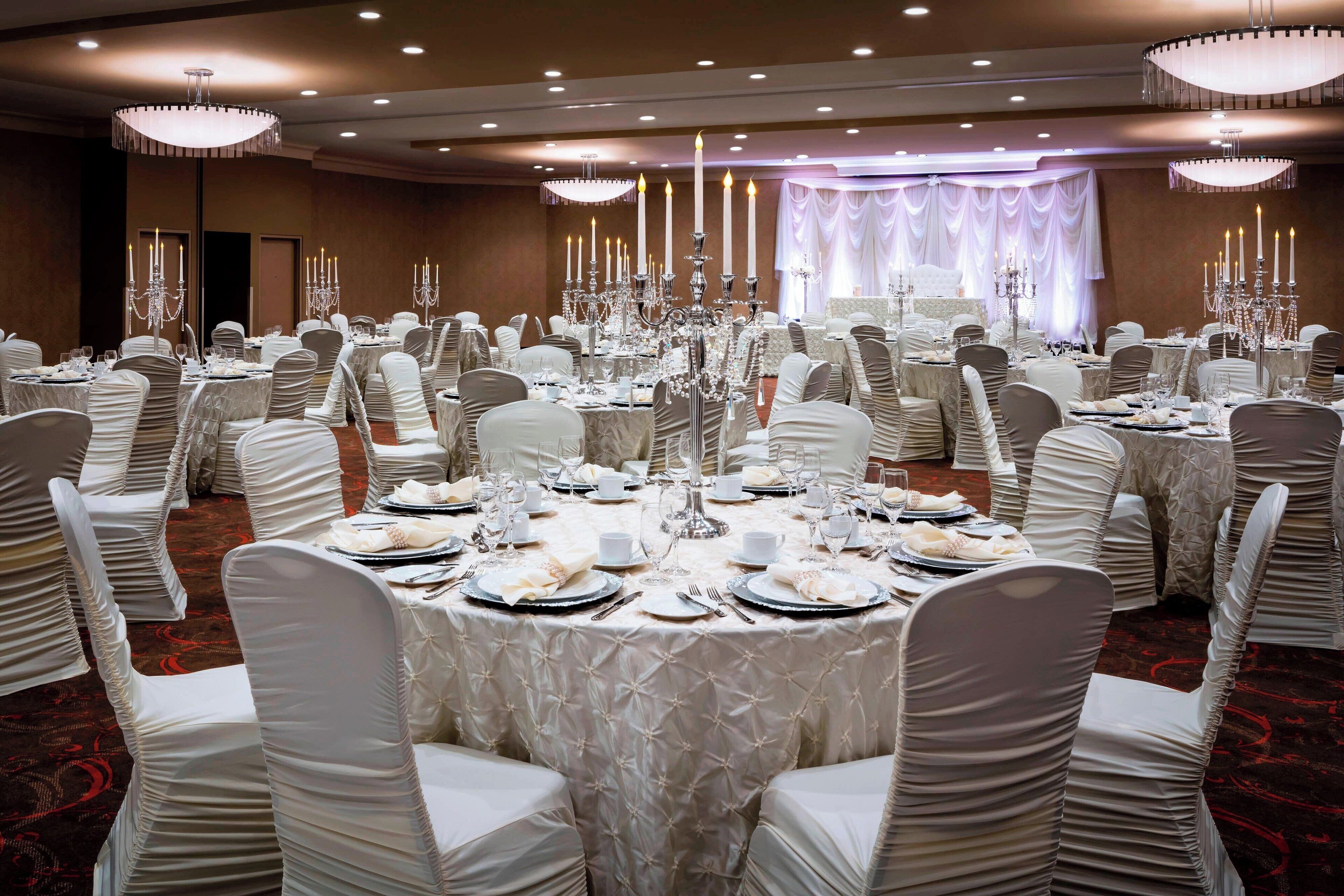 Un salón de banquetes con mesas redondas dispuestas para un evento formal, cada una con mantel blanco, cubertería y candelabros.