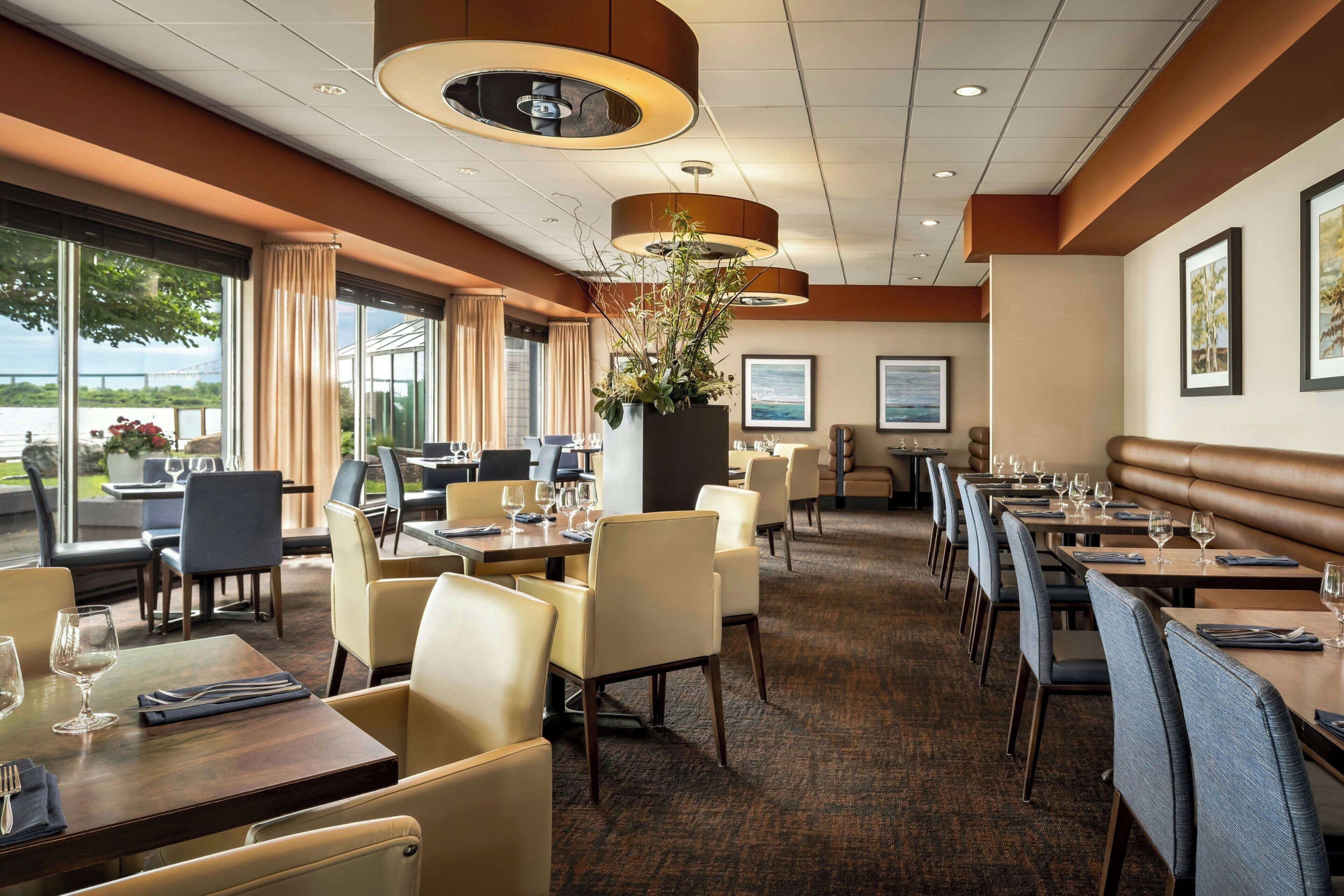 A restaurant with multiple dining tables and chairs, large windows with curtains, and a central plant.