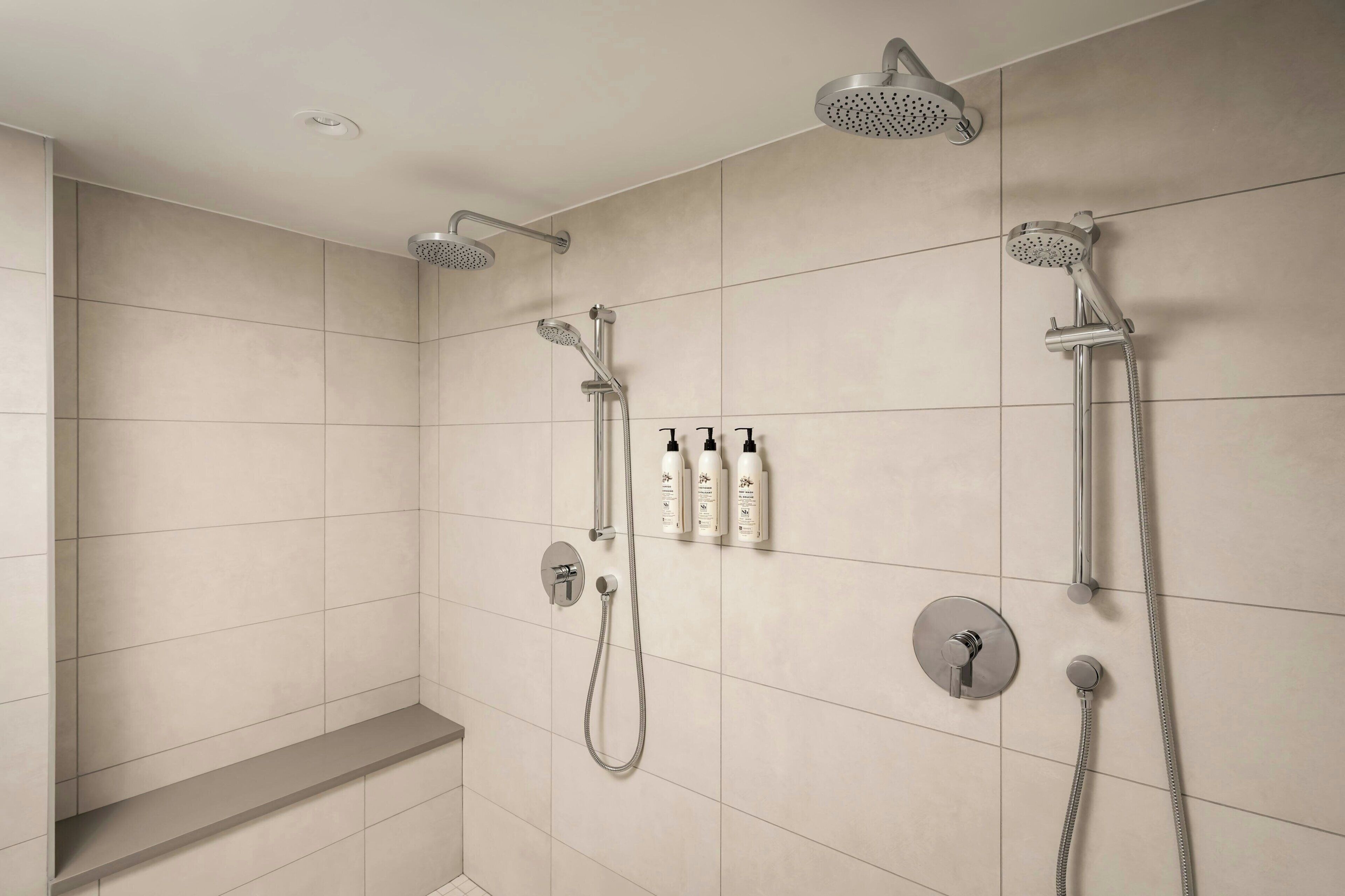 A modern bathroom with a shower area, including a handheld showerhead, a wall-mounted showerhead, and a handheld showerhead with a flexible hose. There are also three bottles of shampoo and conditioner on the wall.