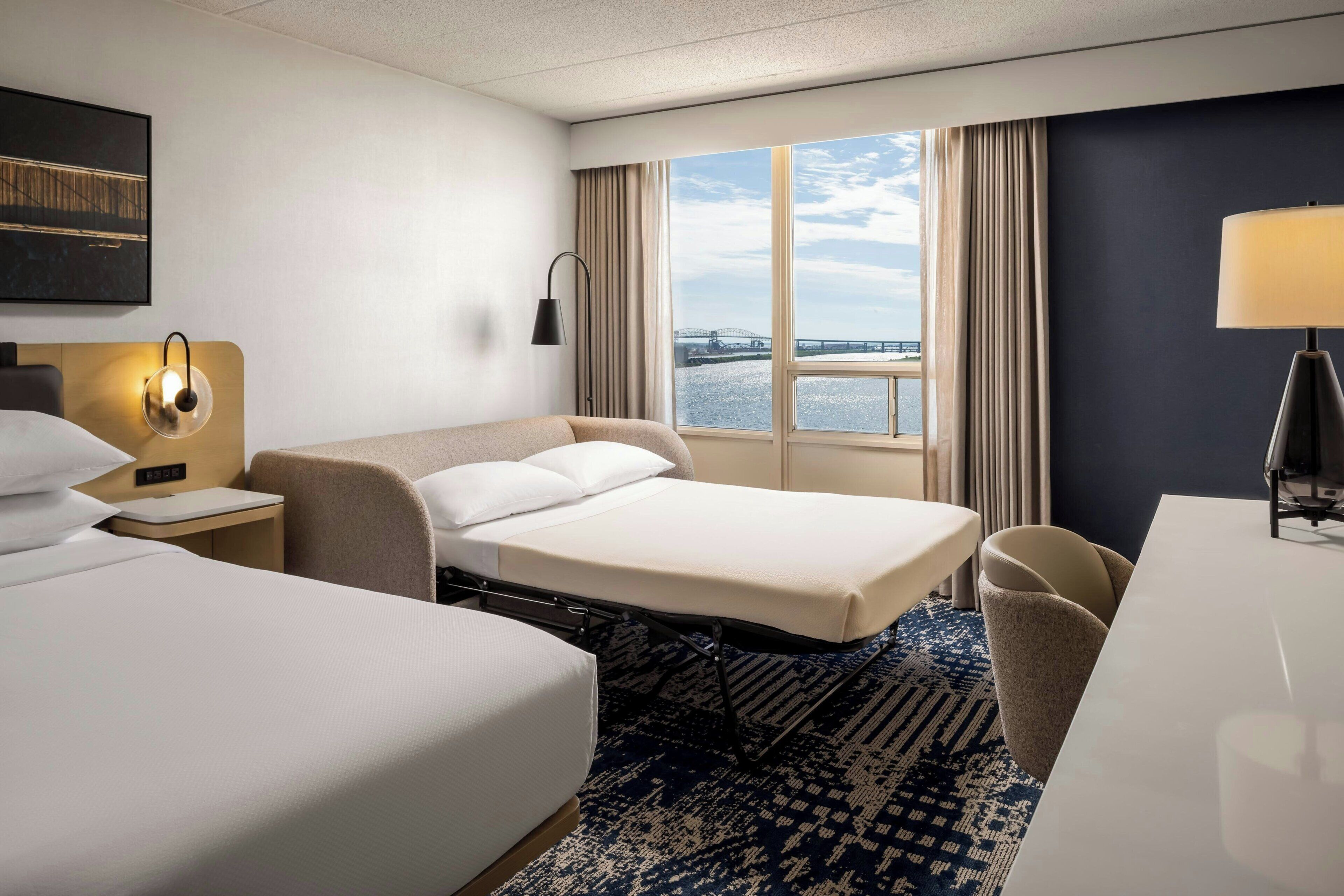 A hotel room with two beds, a desk, a chair, a lamp, and a view of a bridge over water.