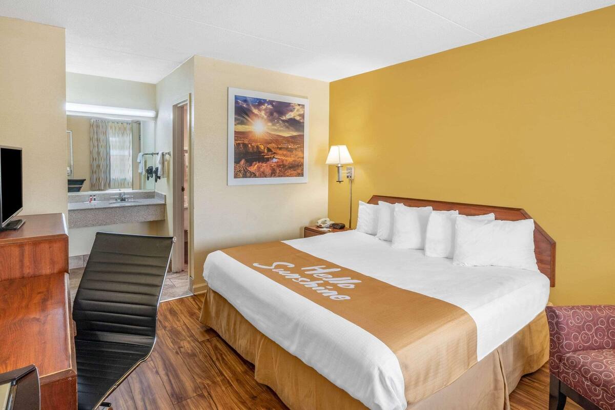 A hotel room with a bed, a desk, a chair, a nightstand, a lamp, and a bathroom visible in the background.