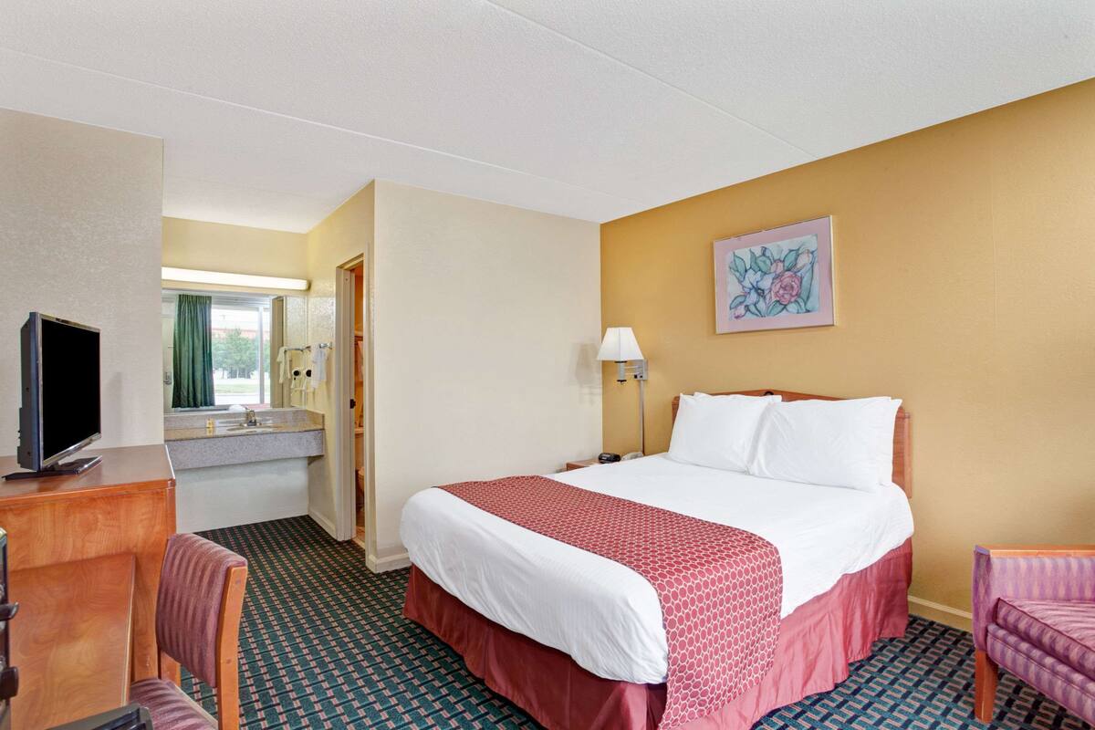 A hotel room with a bed, a desk with a television, a chair, a bathroom with a mirror, and a painting on the wall.