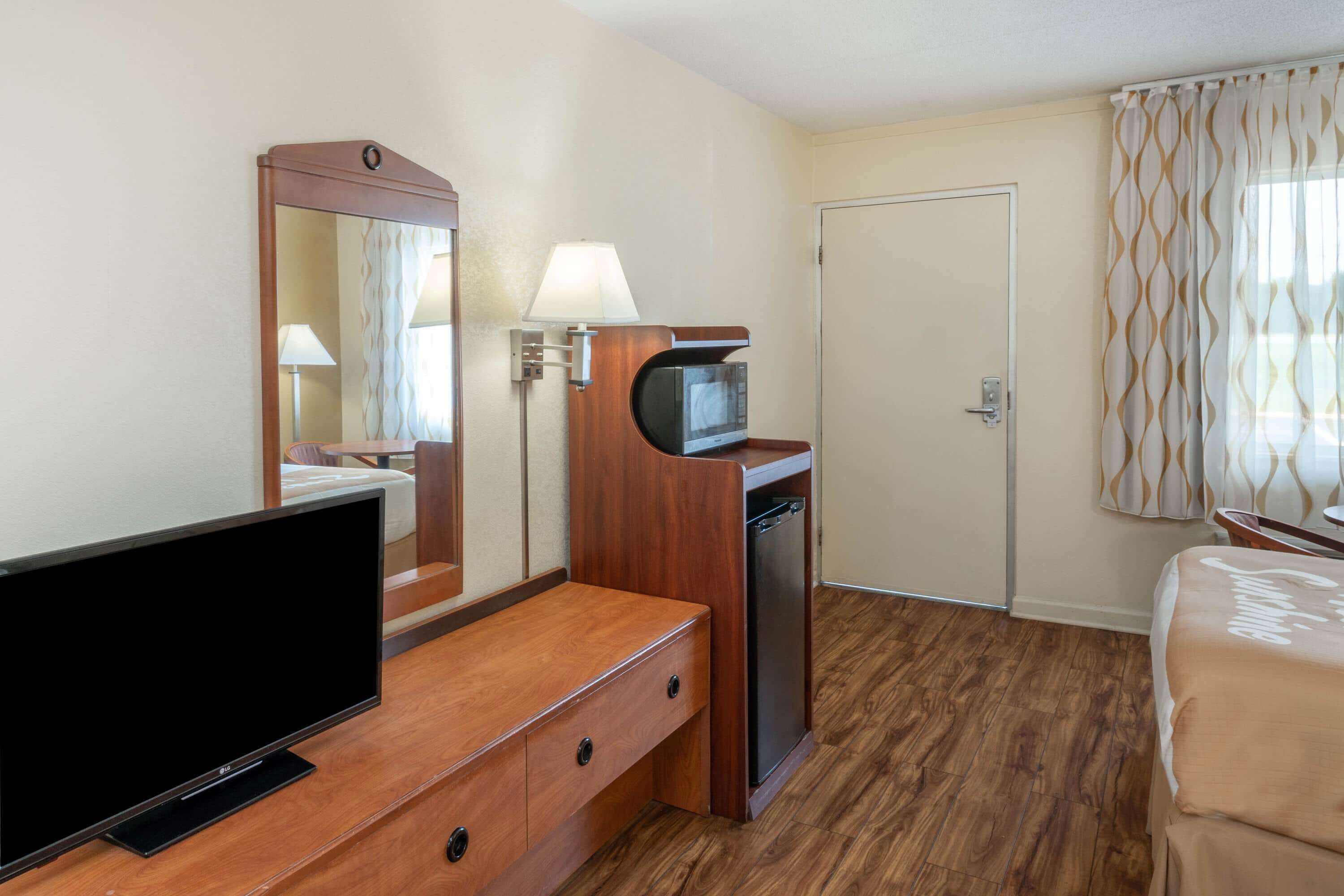 Habitación de hotel con televisor de pantalla plana, escritorio de madera con microondas, lámpara y cama con cubierta color beige.