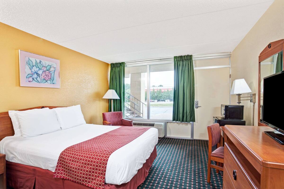 A hotel room with a bed, a desk, a television, a chair, a window with curtains, and a painting on the wall.
