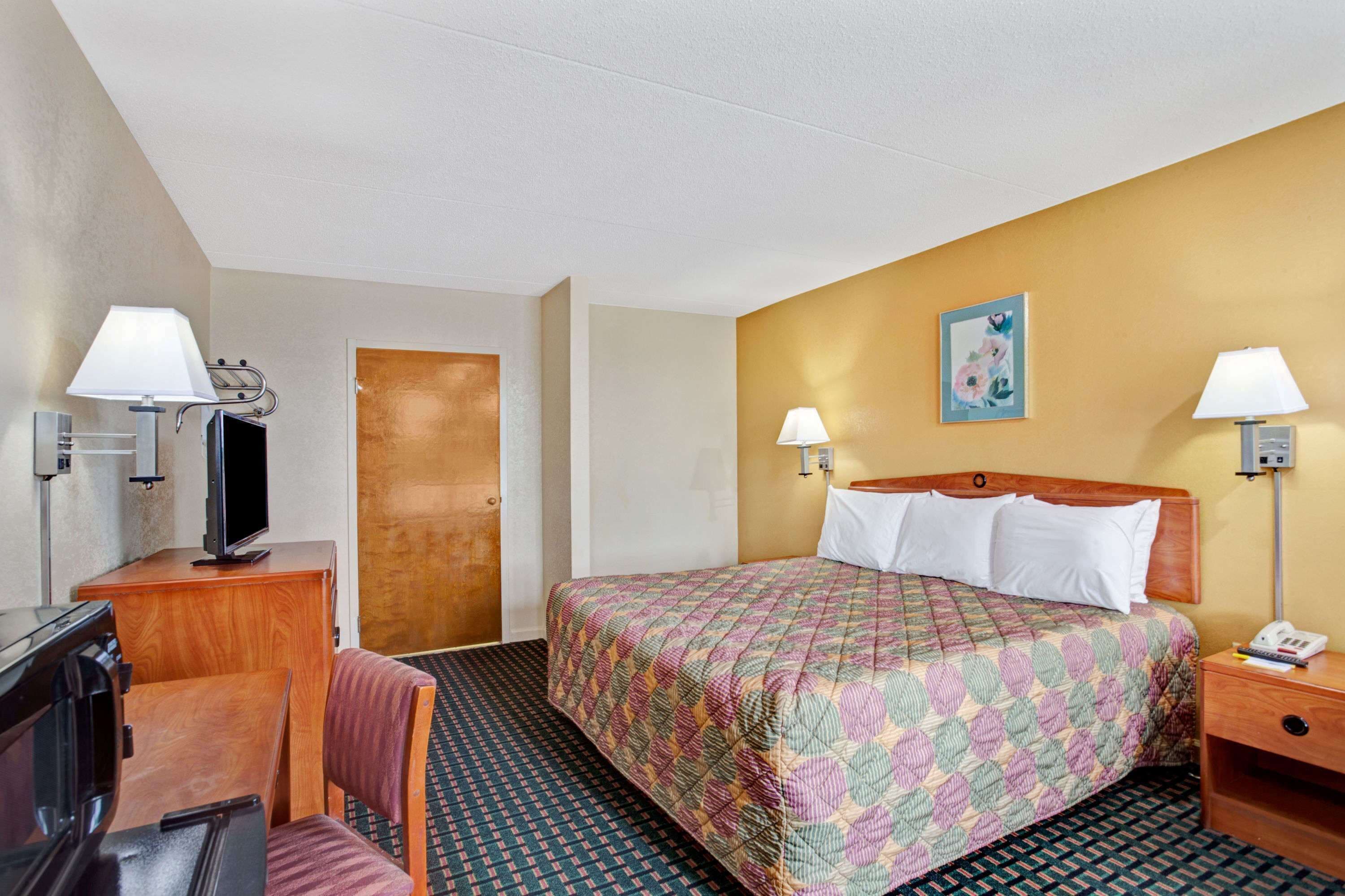 A hotel room with a bed, a desk with a television, two bedside lamps, and a door.