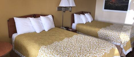 A hotel room with two beds, each with a gold and white floral bedspread, a nightstand, a lamp, and a painting of a sunset on the wall.