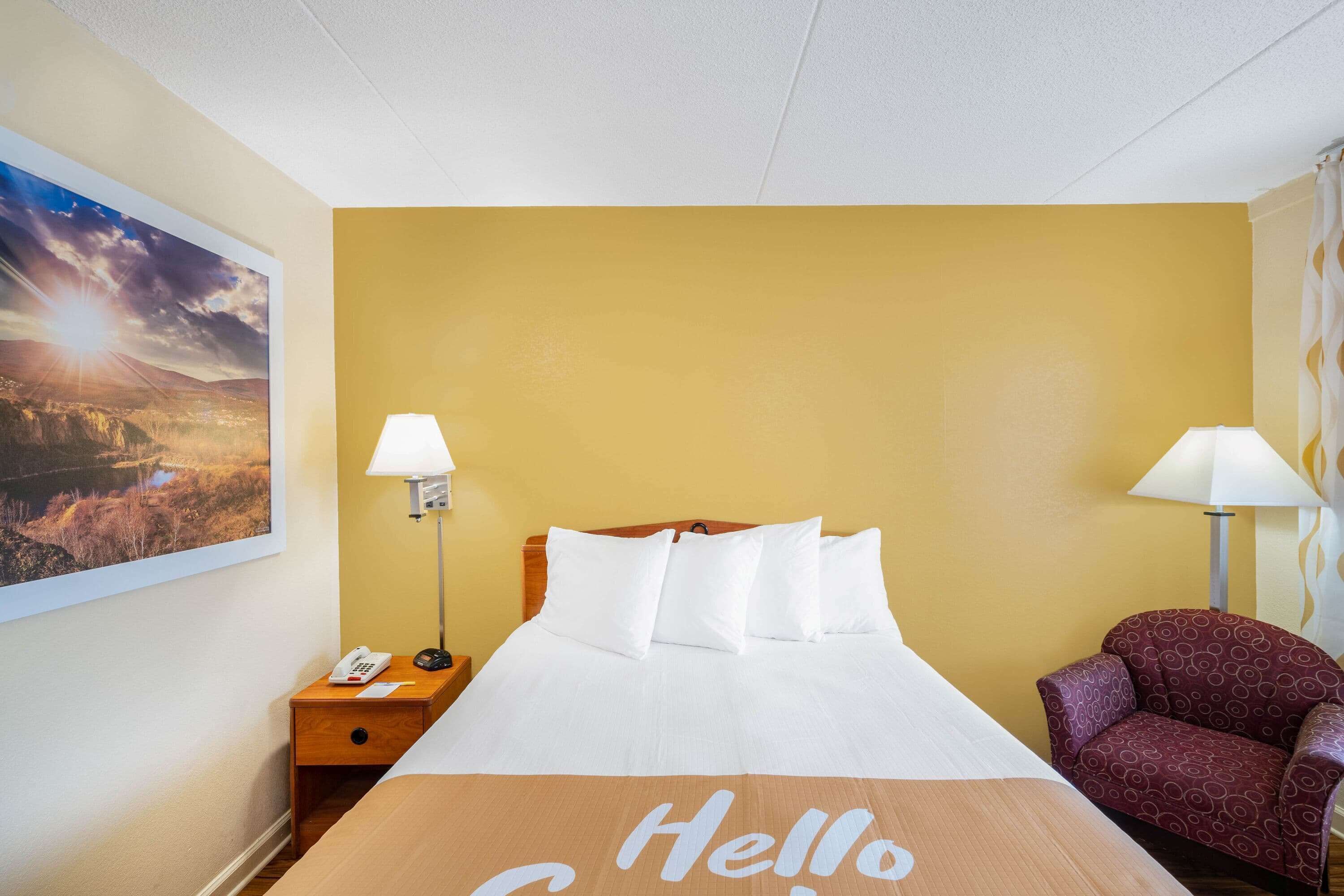 A hotel room with a bed, two bedside lamps, a chair, a nightstand with a phone, and a painting on the wall.