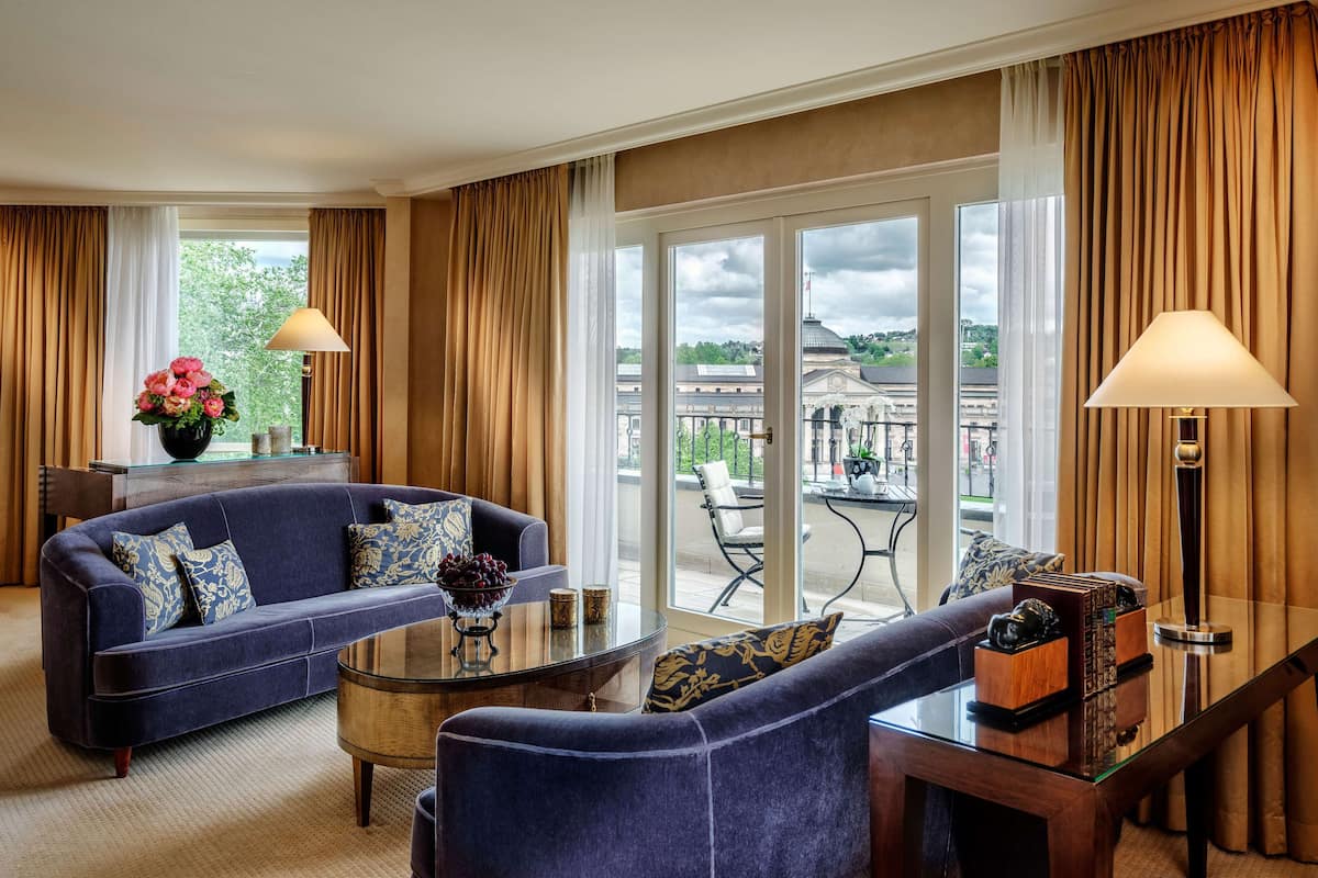 A modern hotel room with a blue sofa, a glass coffee table, and a view of the cityscape through large windows.