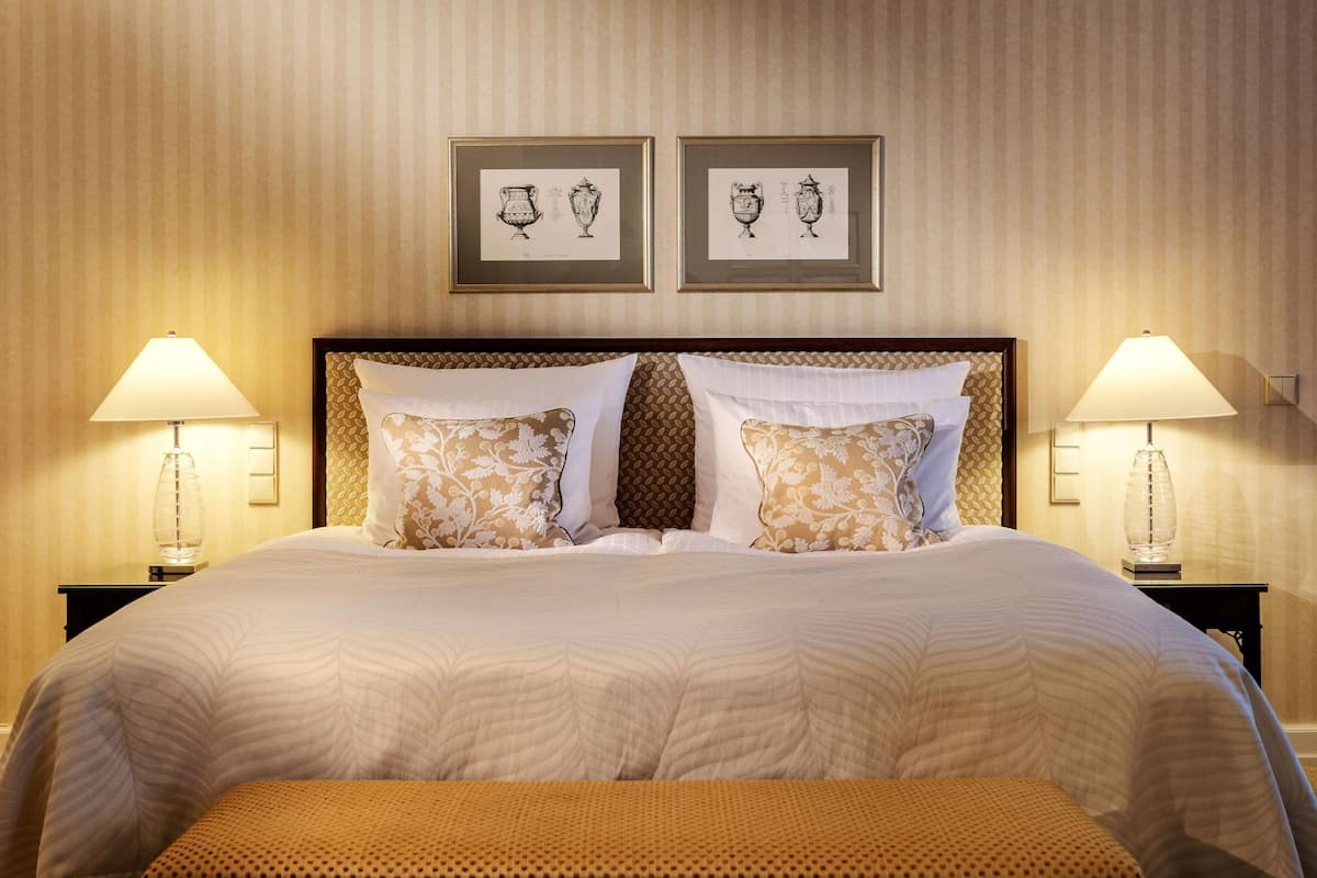 A neatly made bed with a patterned bedspread, two matching pillows, and a bench at the foot of the bed. There are two bedside lamps and two framed pictures above the bed.