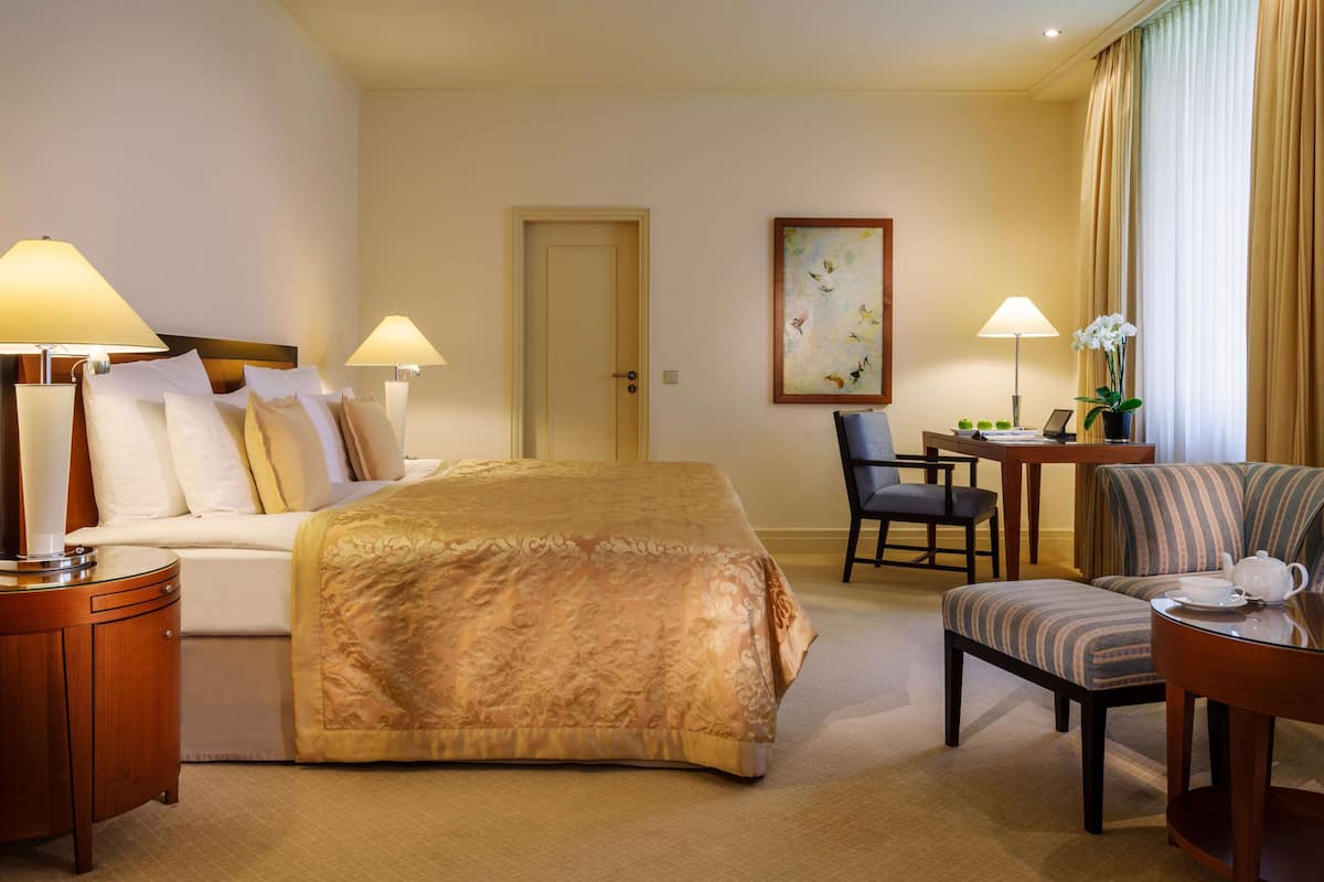 A hotel room with a bed, two chairs, a desk, and a painting on the wall.