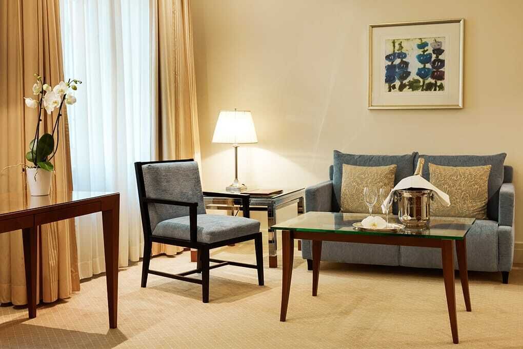 A hotel room with a sofa, armchair, and a glass coffee table. There is a lamp on a side table and a vase with flowers.