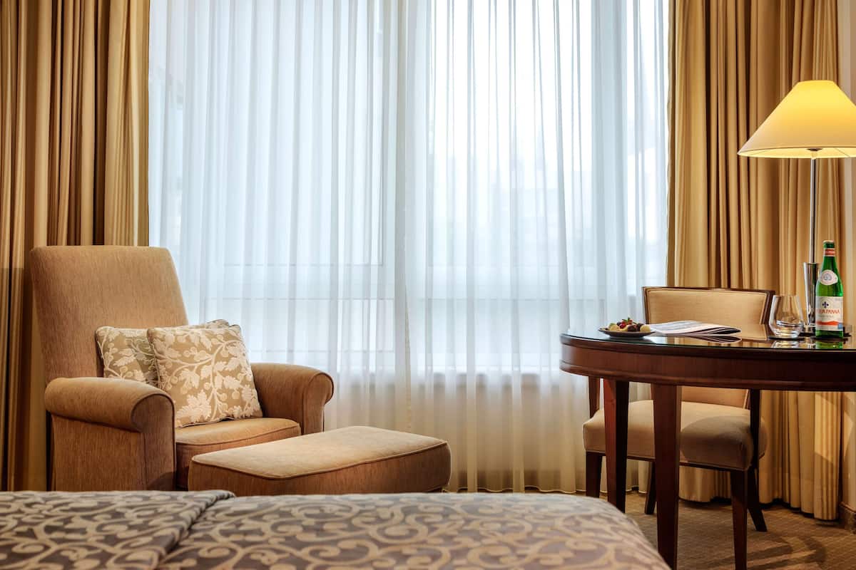 A hotel room with a large bed, a sitting area with a table, and a view through sheer curtains.