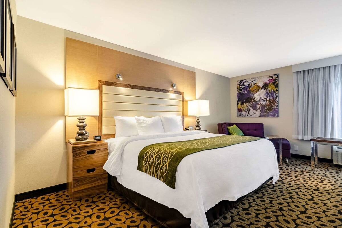 A hotel room with a bed, bedside tables, a desk, a chair, a painting, and a patterned carpet.