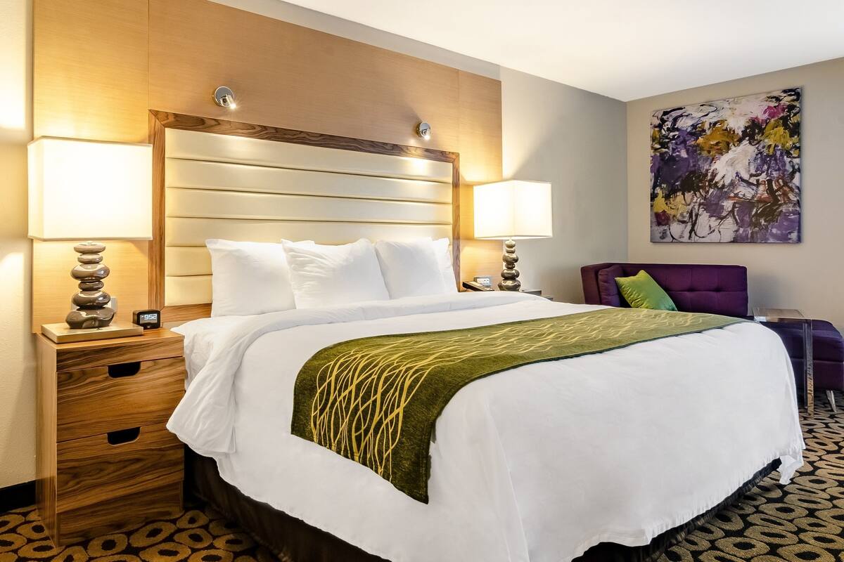 A hotel room with a bed, two bedside tables, a nightstand, a wall-mounted lamp, and a painting.