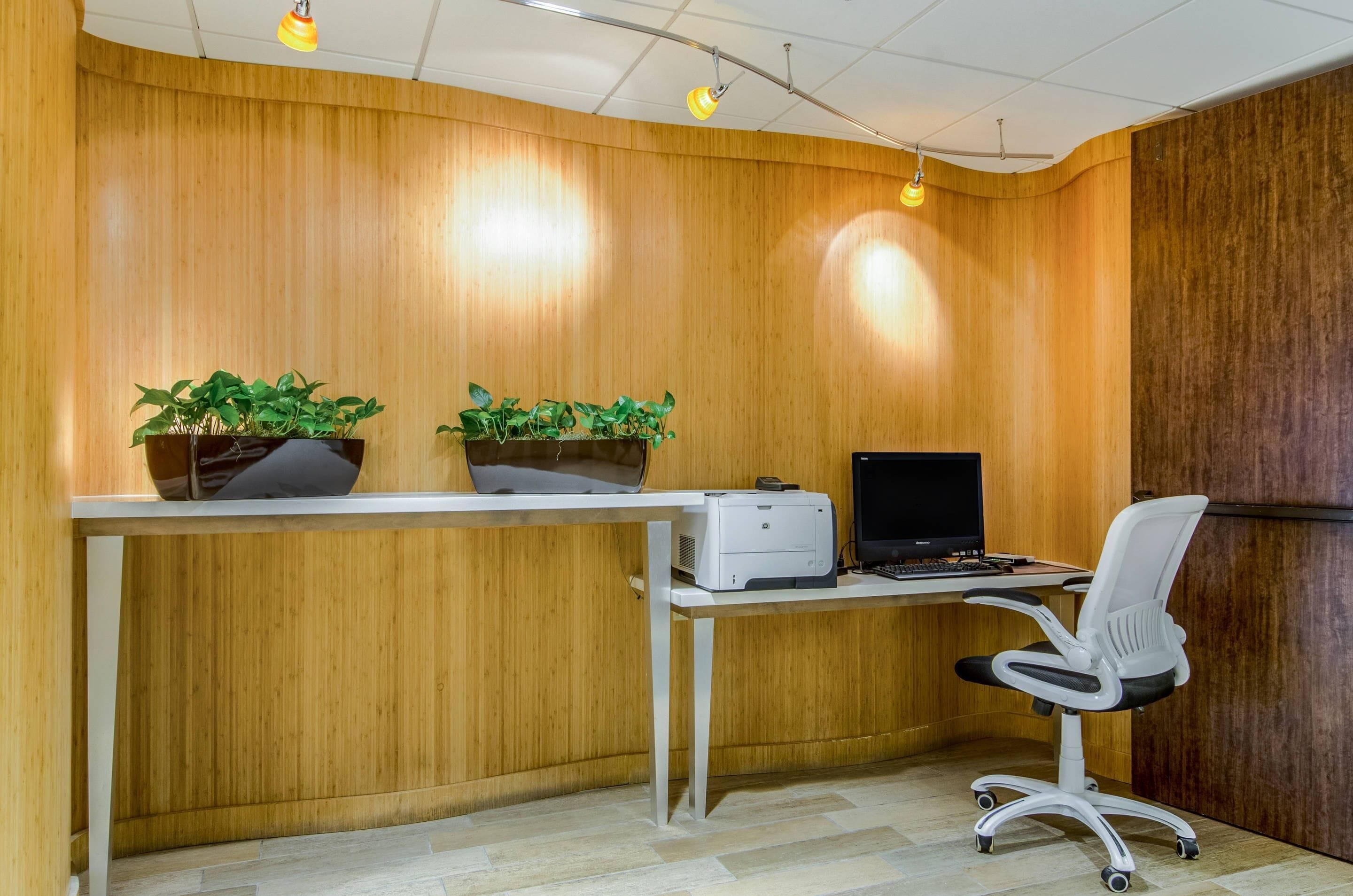 A reception desk with a printer, two potted plants, a computer monitor, and a white office chair.