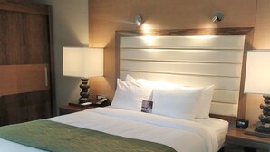 A hotel room with a large bed, two bedside tables, a nightstand, a headboard, and a wall-mounted lamp.