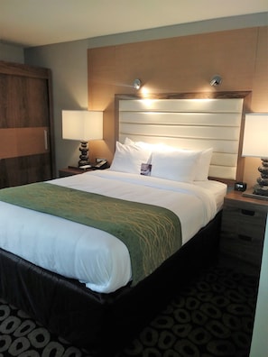 Une chambre d’hôtel avec un grand lit, deux tables de chevet, une petite table, une tête de lit et une lampe fixée au mur.