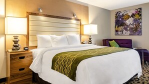 A hotel room with a large bed, two bedside tables, a nightstand, a wall-mounted lamp, a decorative pillow, and a painting on the wall.