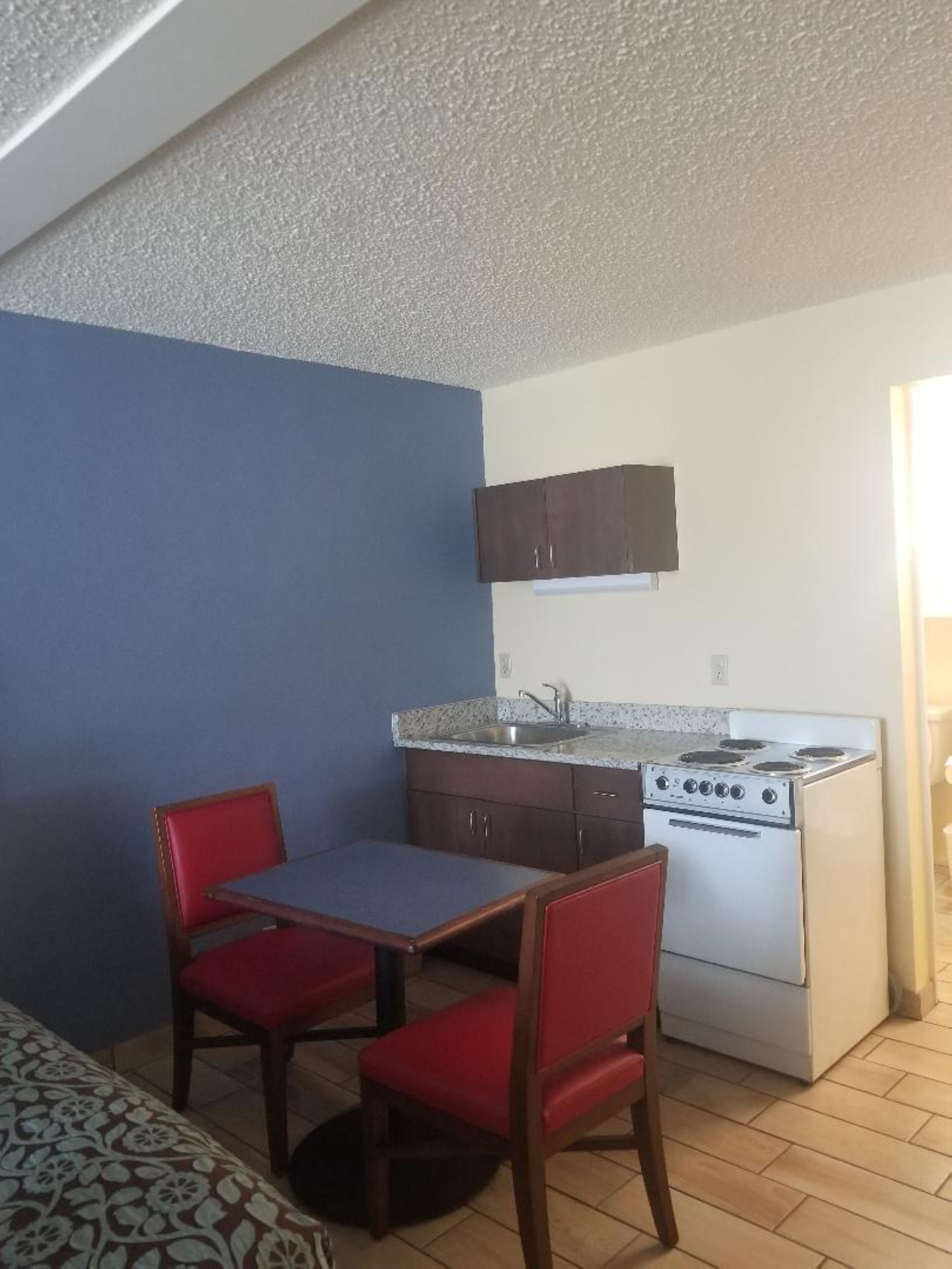 Una cocina pequeña y funcional con estufa, lavamanos y dos sillas rojas junto a una mesa.