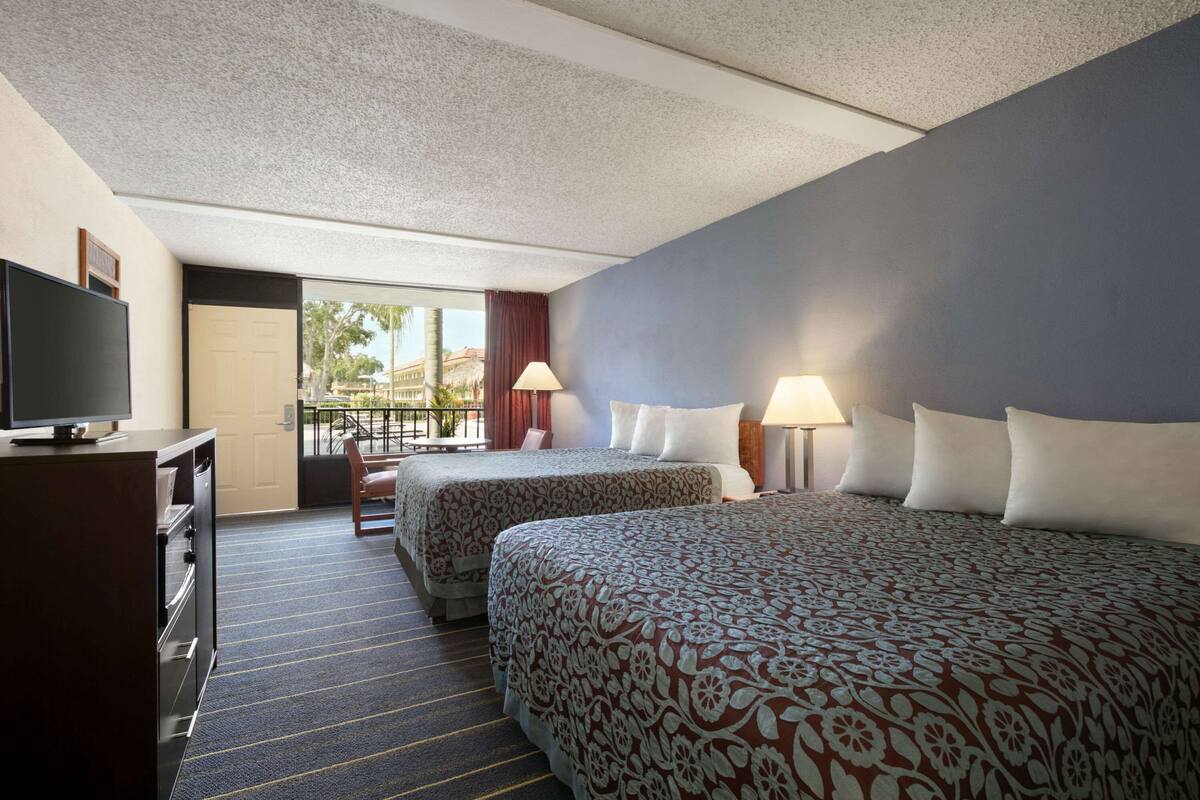 Ein Hotelzimmer mit zwei Betten, einem Fernseher, einem Schreibtisch und Blick auf Palmen.