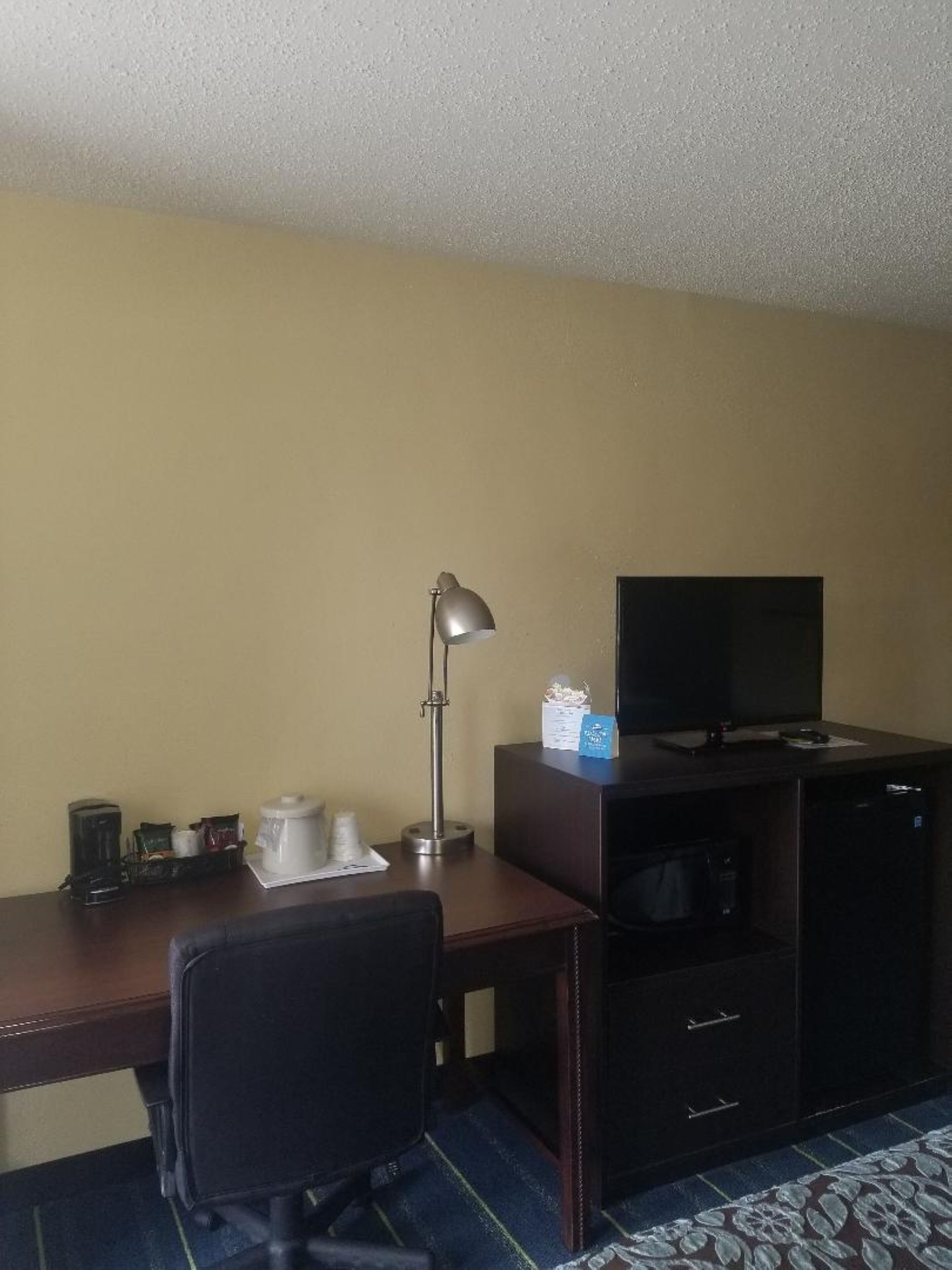 Een kleine hotelkamer met een bureau, een stoel, een televisie, een koffiezetapparaat en een magnetron.