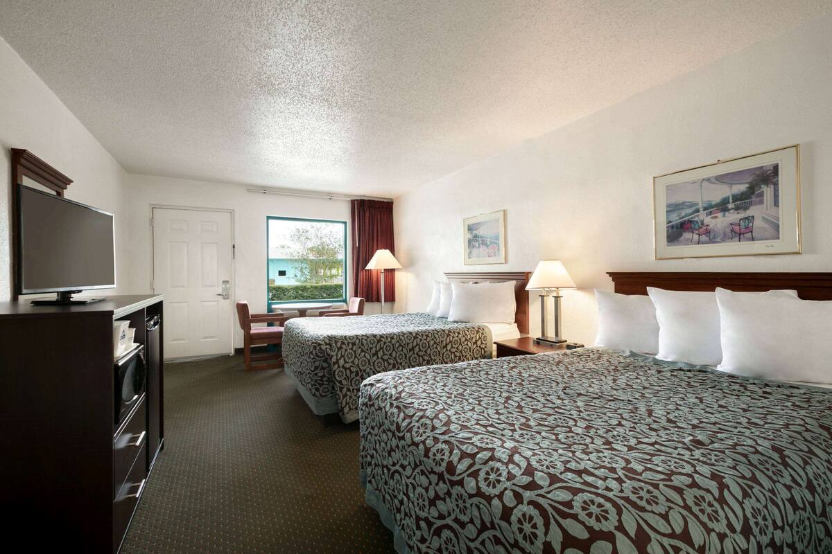 Ein Hotelzimmer mit zwei Betten, einem Fernseher, einem Schreibtisch und einem Fenster mit Blick ins Freie.