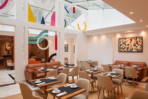 A modern dining area with high ceilings, geometric wall art, and a mix of seating options including tables and sofas.