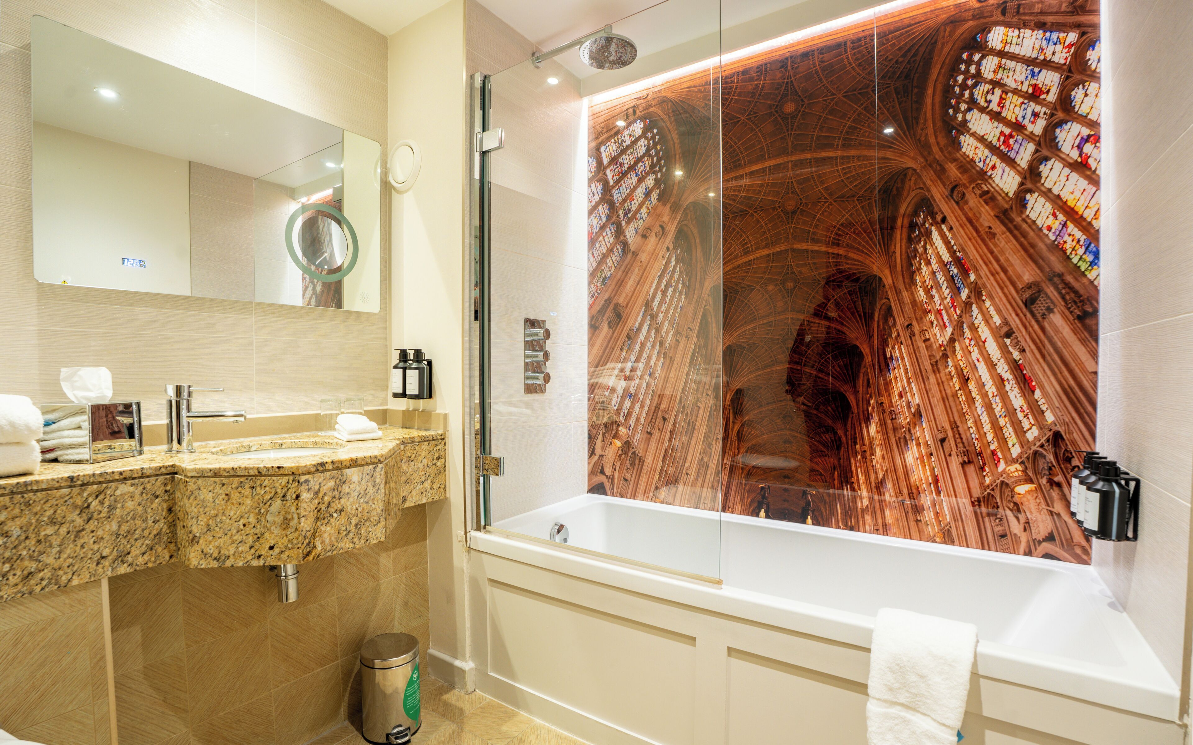 A modern bathroom with a large mirror, granite countertop, and a bathtub with a stained glass wall.