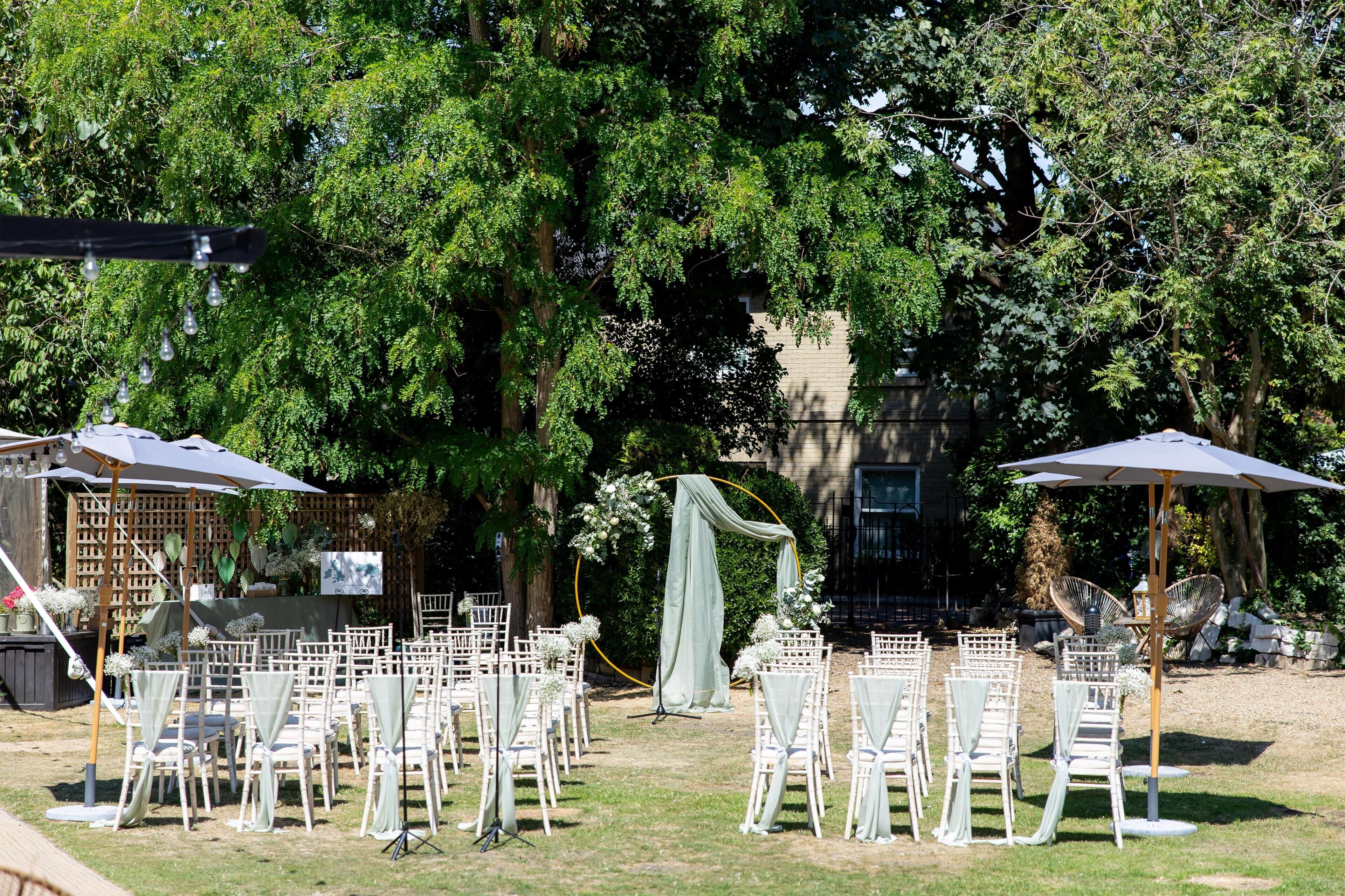 An outdoor wedding setup with white chairs and tables, a draped arch, and a backdrop of trees and a building.