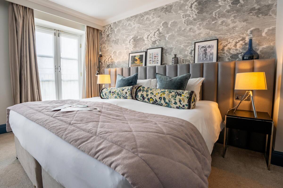 A hotel room with a large bed, a headboard with framed pictures, a nightstand with a lamp, and a window with curtains.