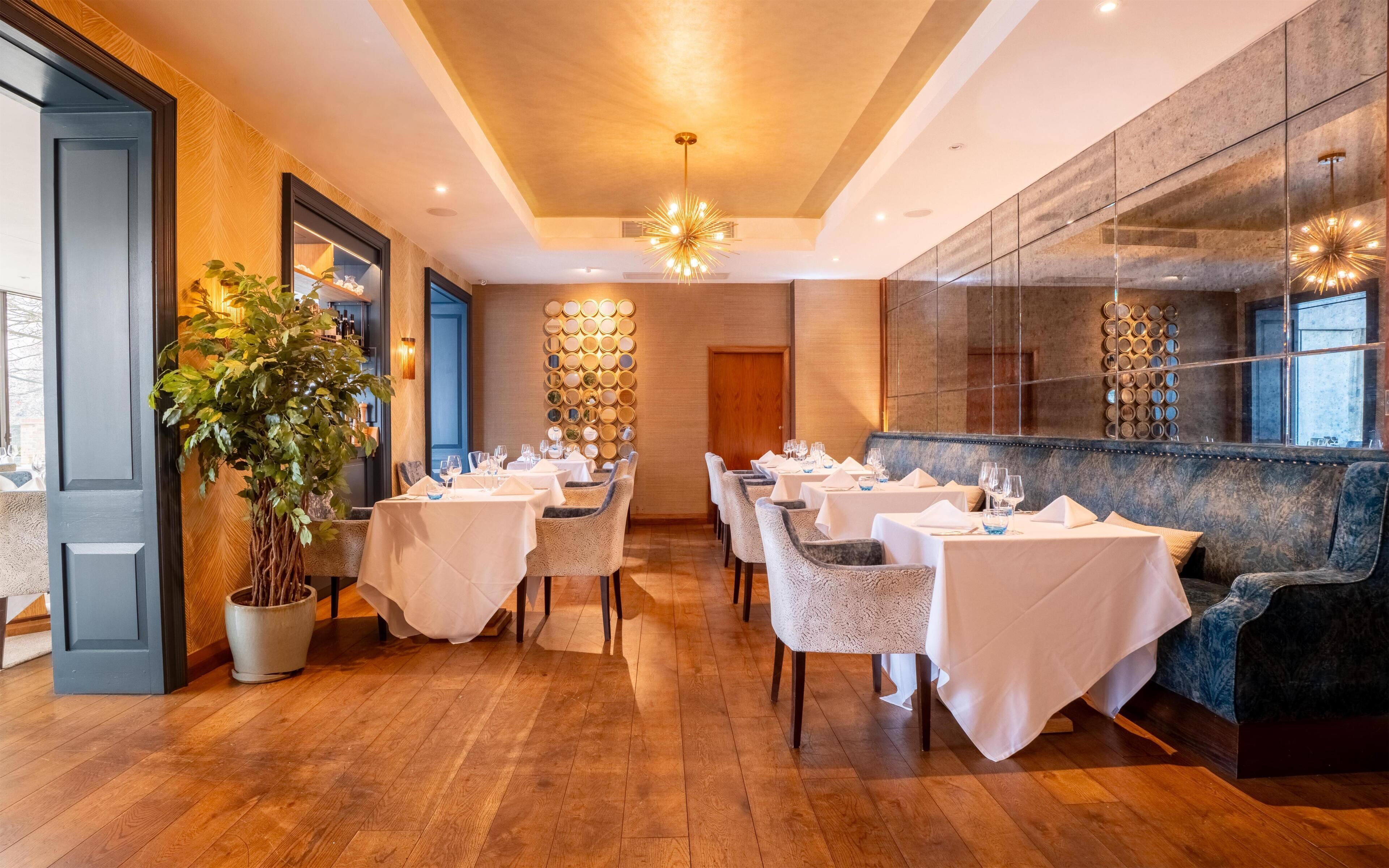 A restaurant with wooden flooring, white tablecloths, and a large potted plant.