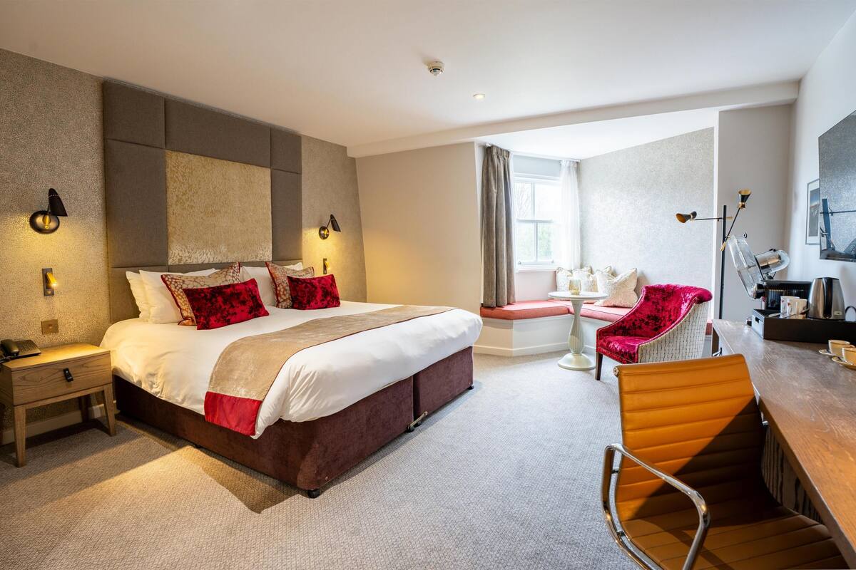 A hotel room with a bed, bedside tables, a desk with a chair, a coffee machine, and a window with curtains.