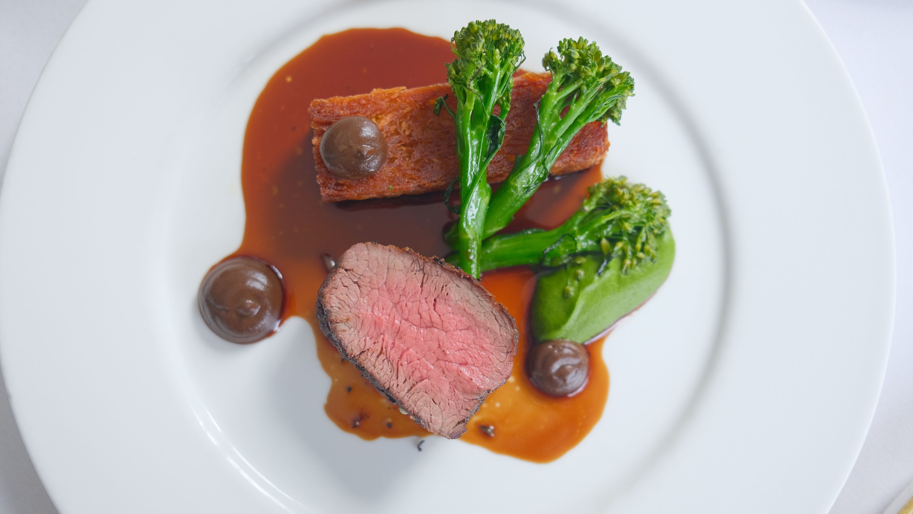 A plate with a slice of meat, broccoli, and two round objects, possibly sauces, on a white plate.