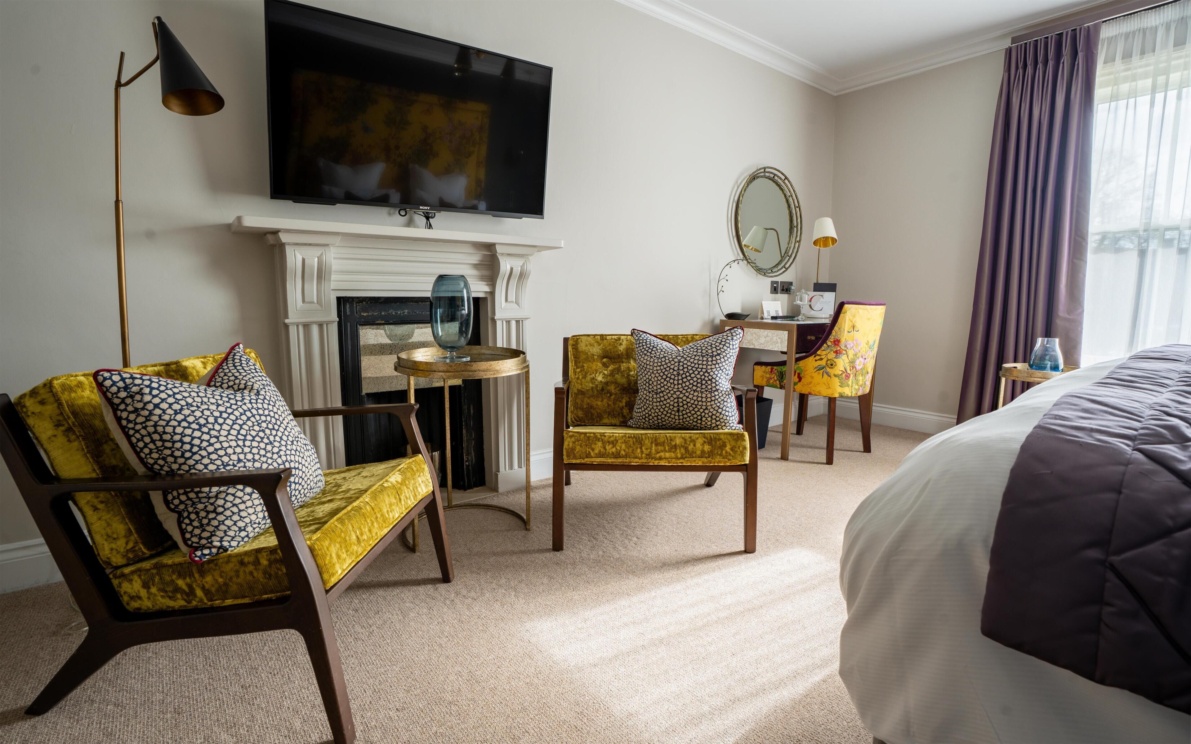 A hotel room with a flat-screen TV, a fireplace, two armchairs, a round table, a bed, and a window with curtains.