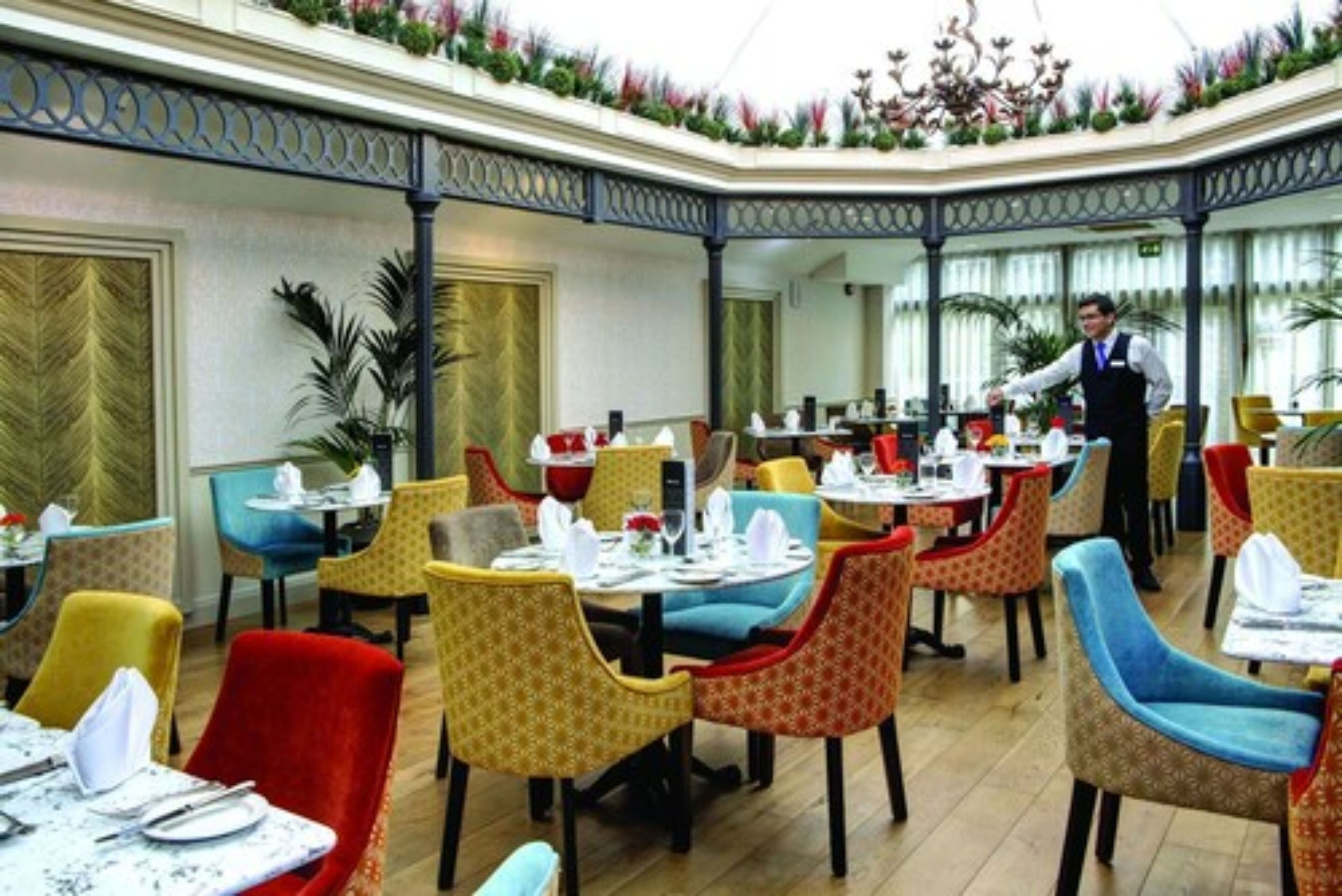 A restaurant with colorful chairs and tables, a waiter, and a decorative ceiling.