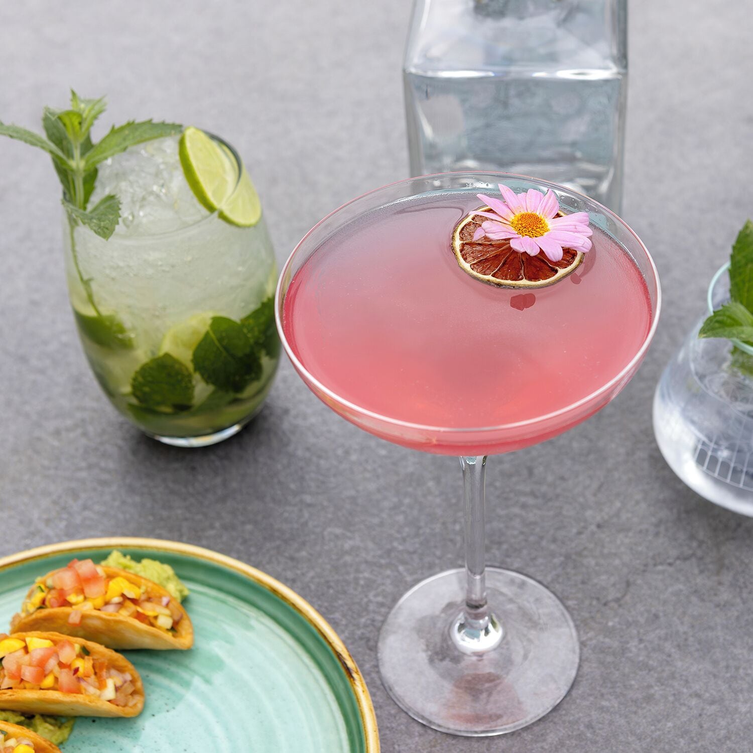 Een cocktailglas met een roze drankje, een glas bevroren drank met een limoen en munt, en een bord met taco's.