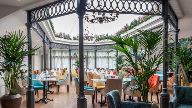 A spacious indoor dining area with multiple tables and chairs, a central decorative column, and potted plants. - The Gonville Hotel, a member of Radisson Individuals (Cambridge)