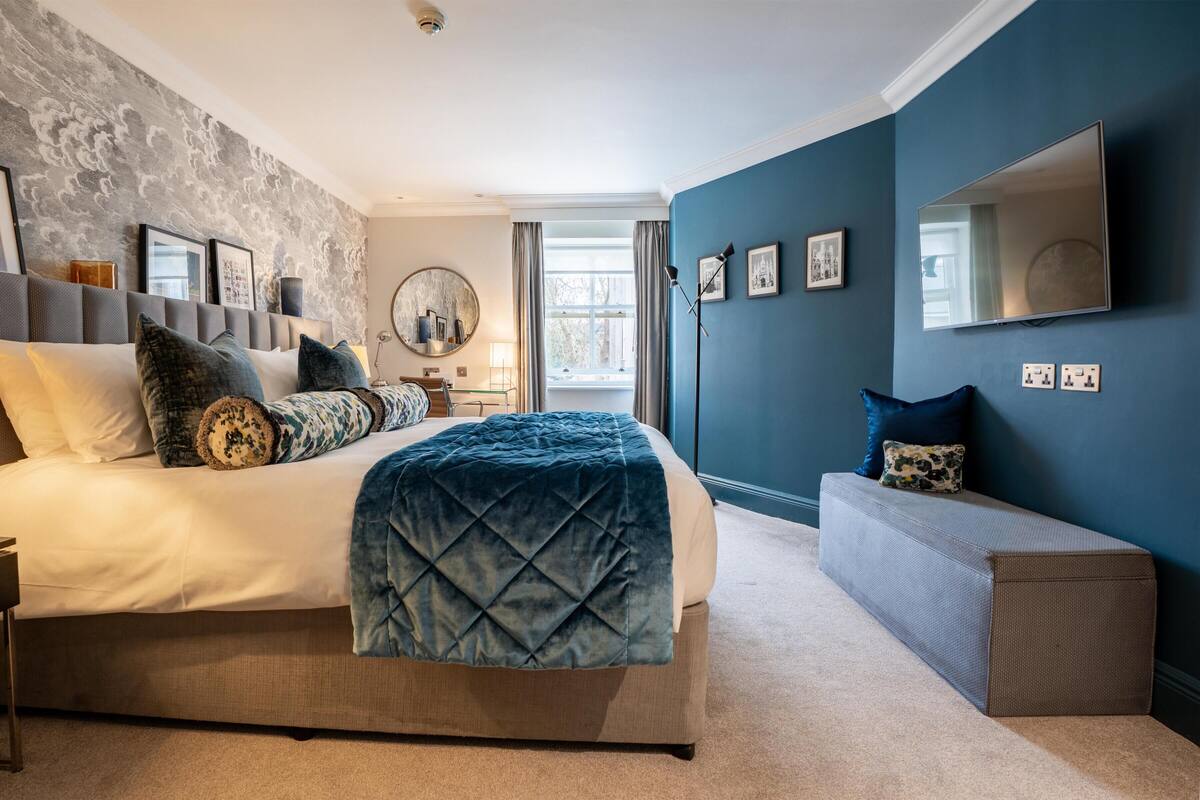 A modern bedroom with a large bed, a bedside table, a desk, a chair, a mirror, and a window with curtains.