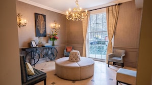 A room with a round ottoman, a chair, a desk with a bicycle, a chandelier, and a window with curtains. - The Gonville Hotel, a member of Radisson Individuals (Cambridge)