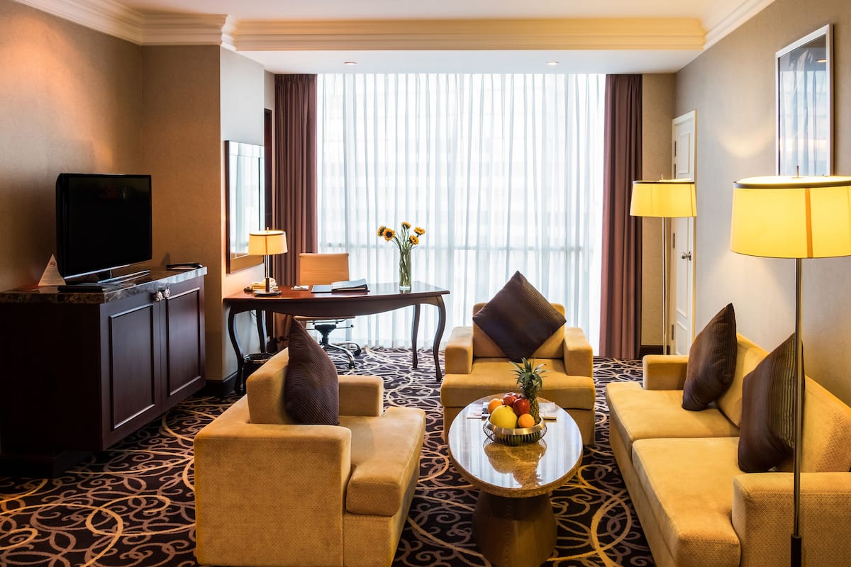 Habitación de hotel con sofá, sillones, una mesa de centro con fruta, televisor, escritorio y una ventana grande con cortinas.