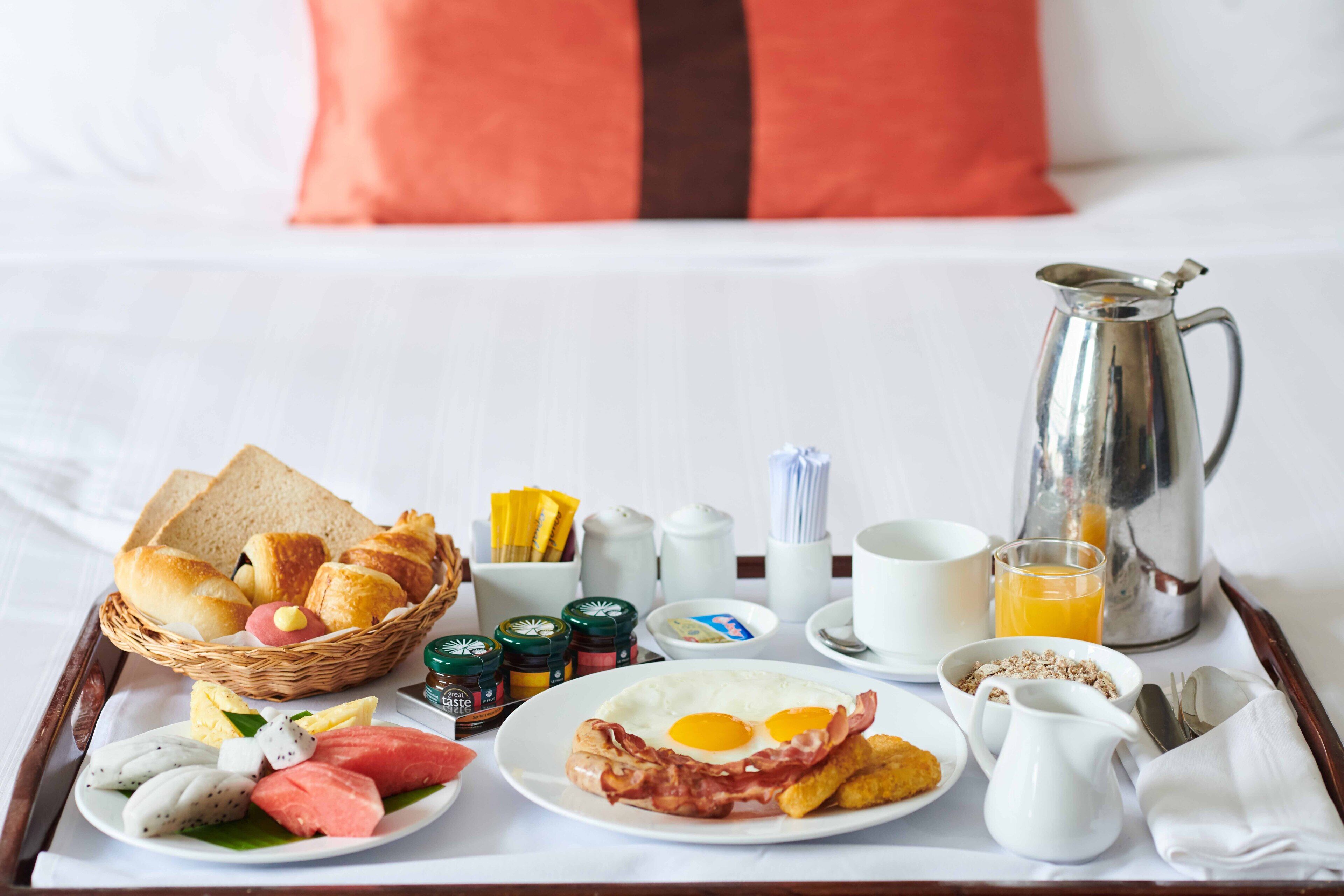 A breakfast tray with toast, eggs, bacon, and fruit on a hotel bed.