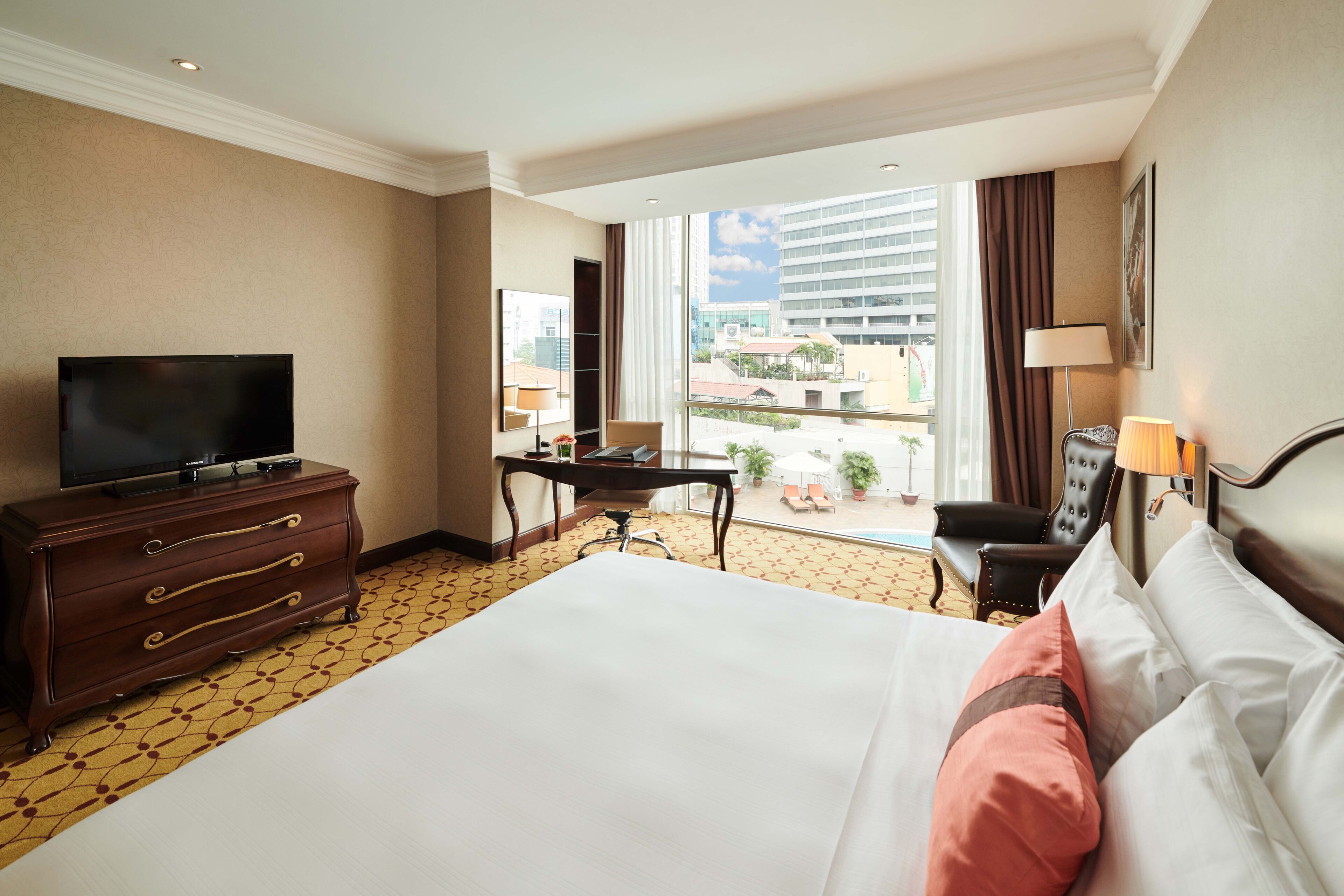 A hotel room with a large bed, a desk, a television, and a view of the city through the window.