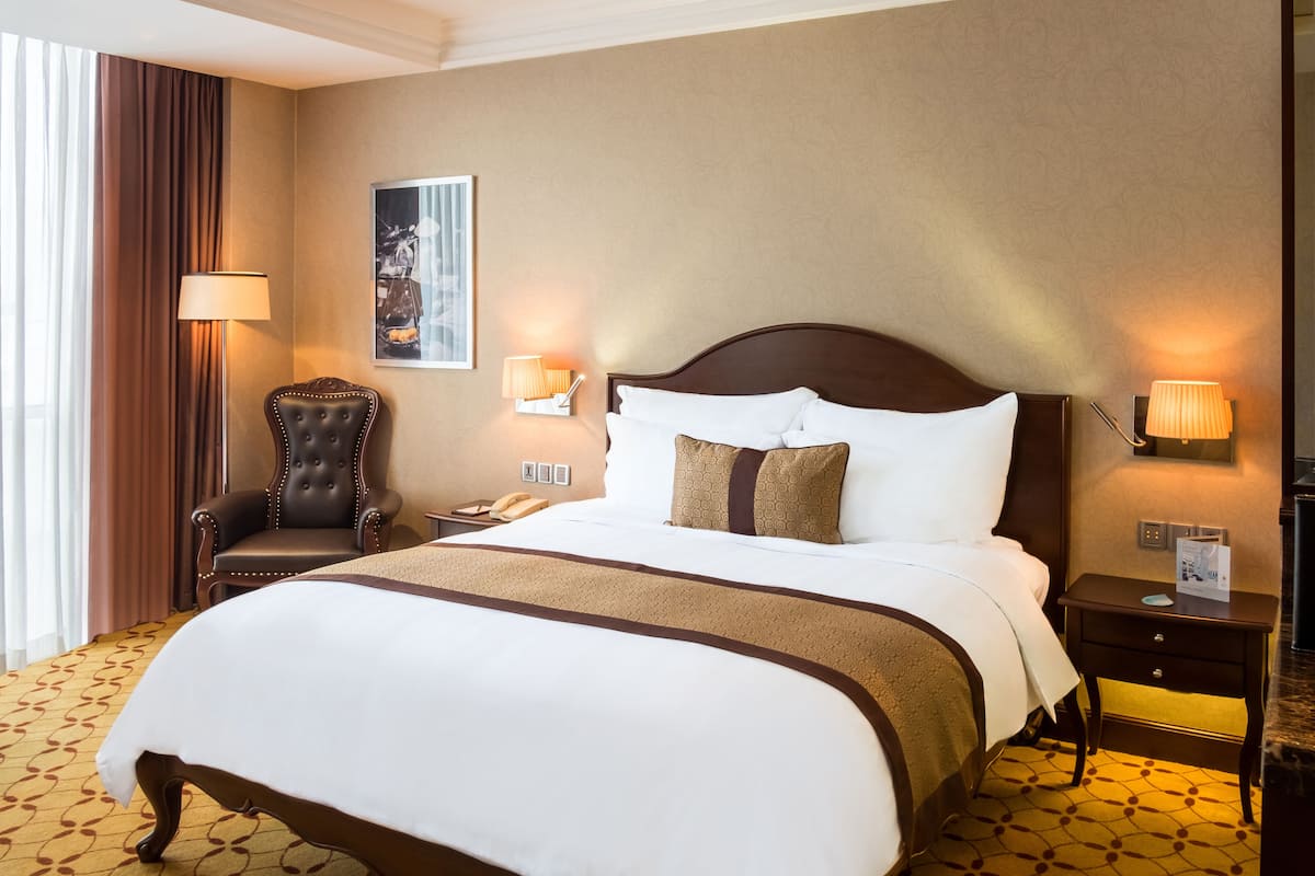 Habitación de hotel con una cama grande, una silla, dos lámparas de noche, una mesita de noche y un cuadro en la pared.