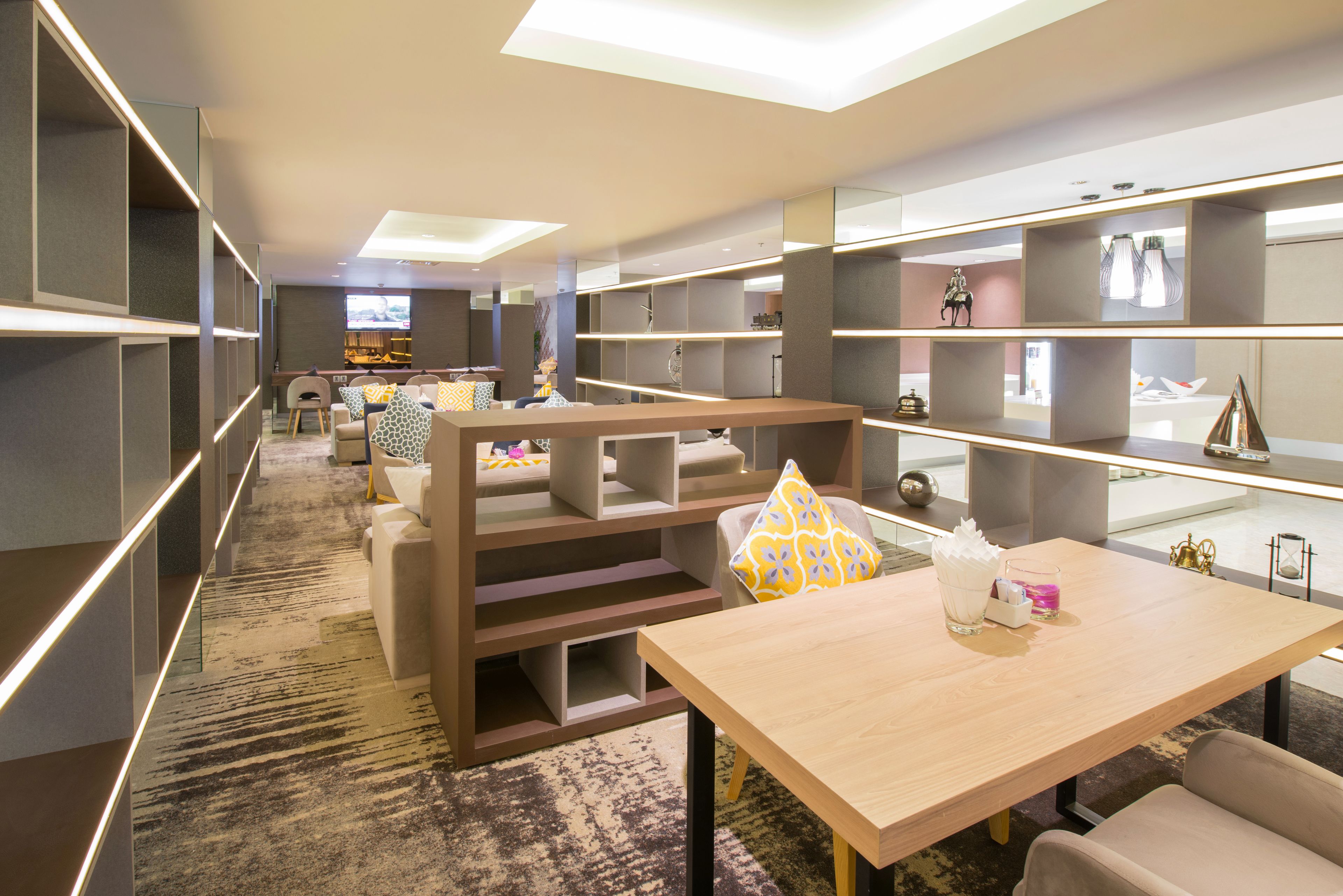 A modern hotel room with a large bed, a desk, and a TV. There are shelves with decorative items and a carpeted floor.